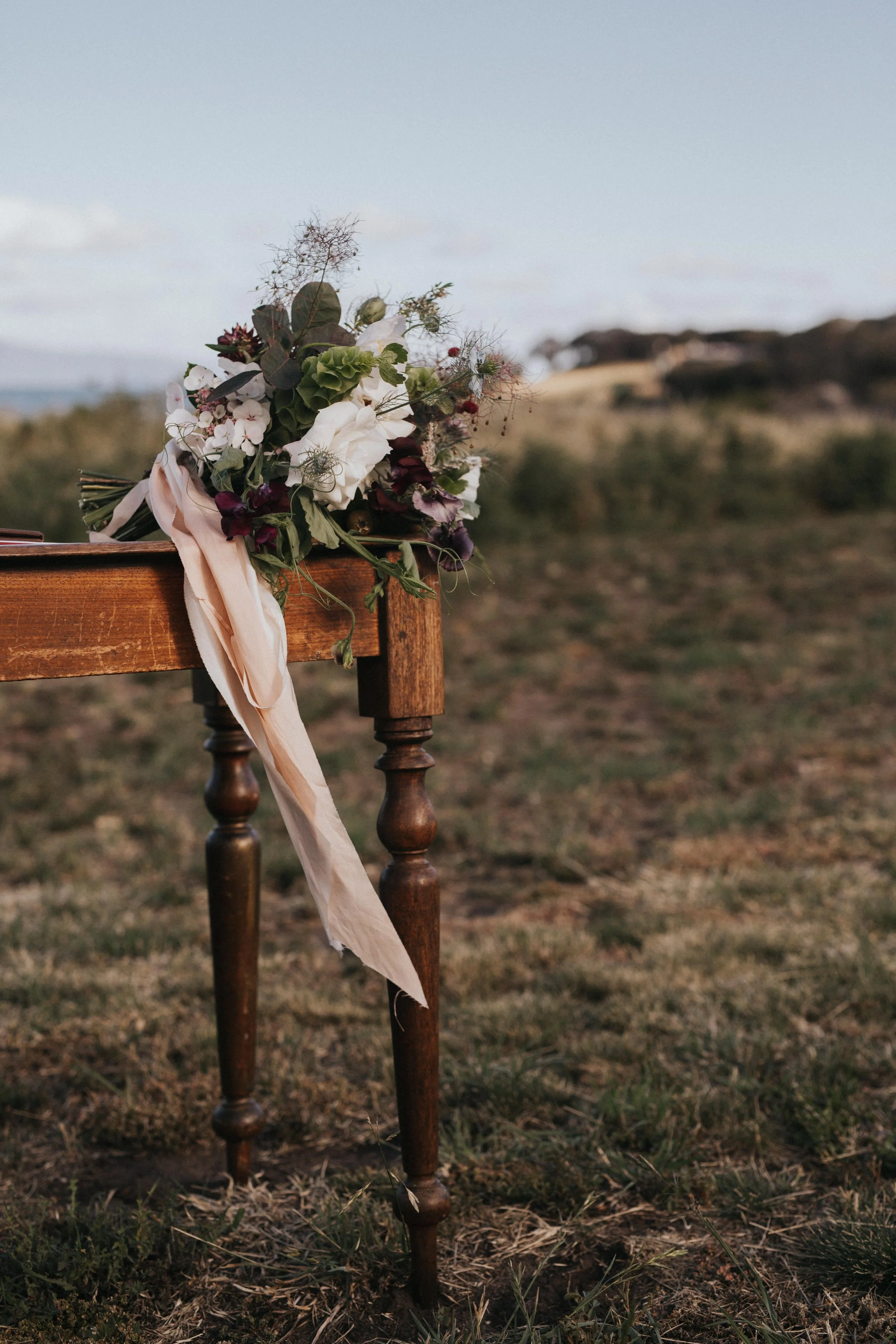 A floral arrangement with white and purple flowers and green foliage, with a light pink fabric ribbon, resting on a wooden table outdoors in a grassy field.