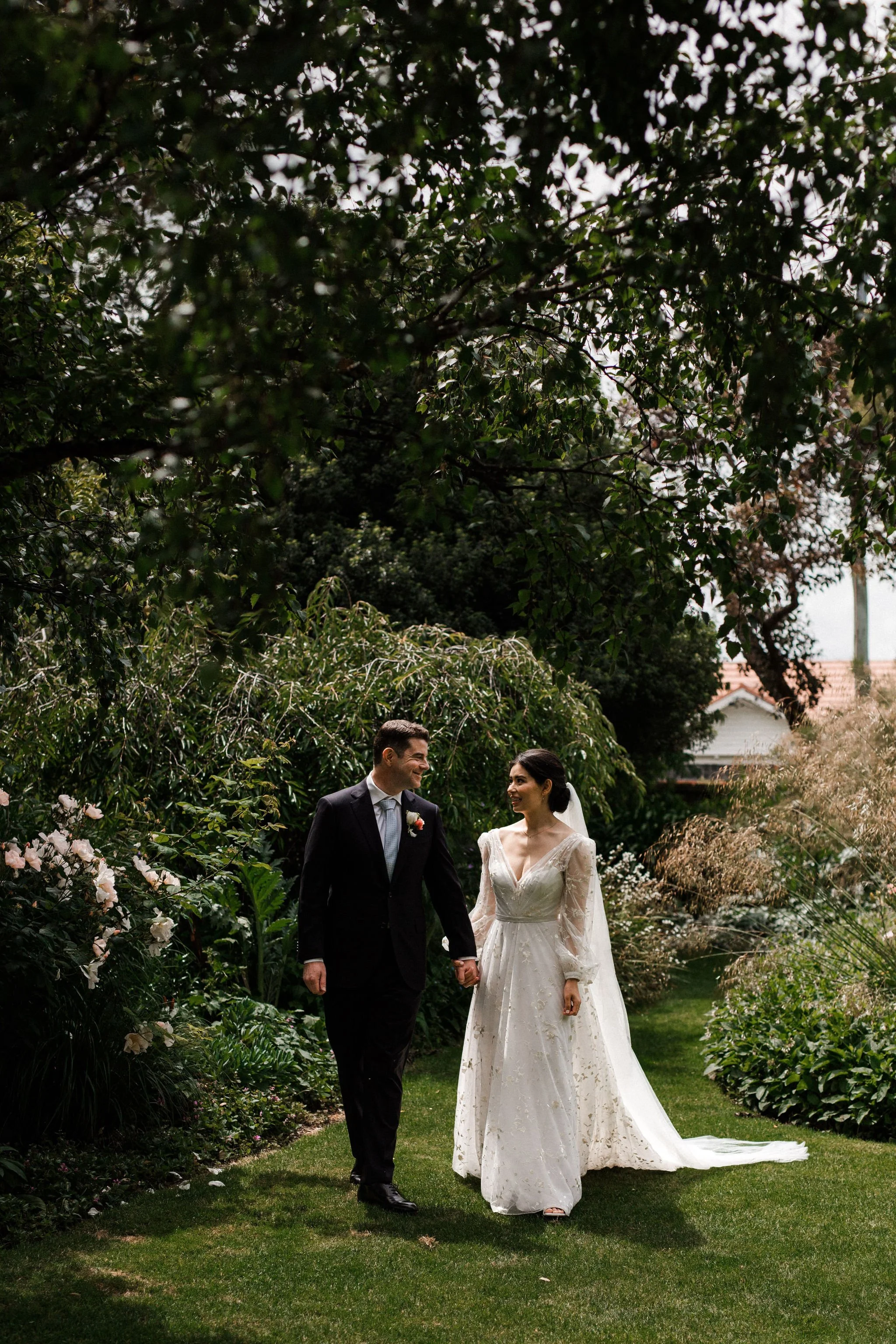 A bride and groom holding hands, walking in a lush garden, smiling at each other during their wedding day.