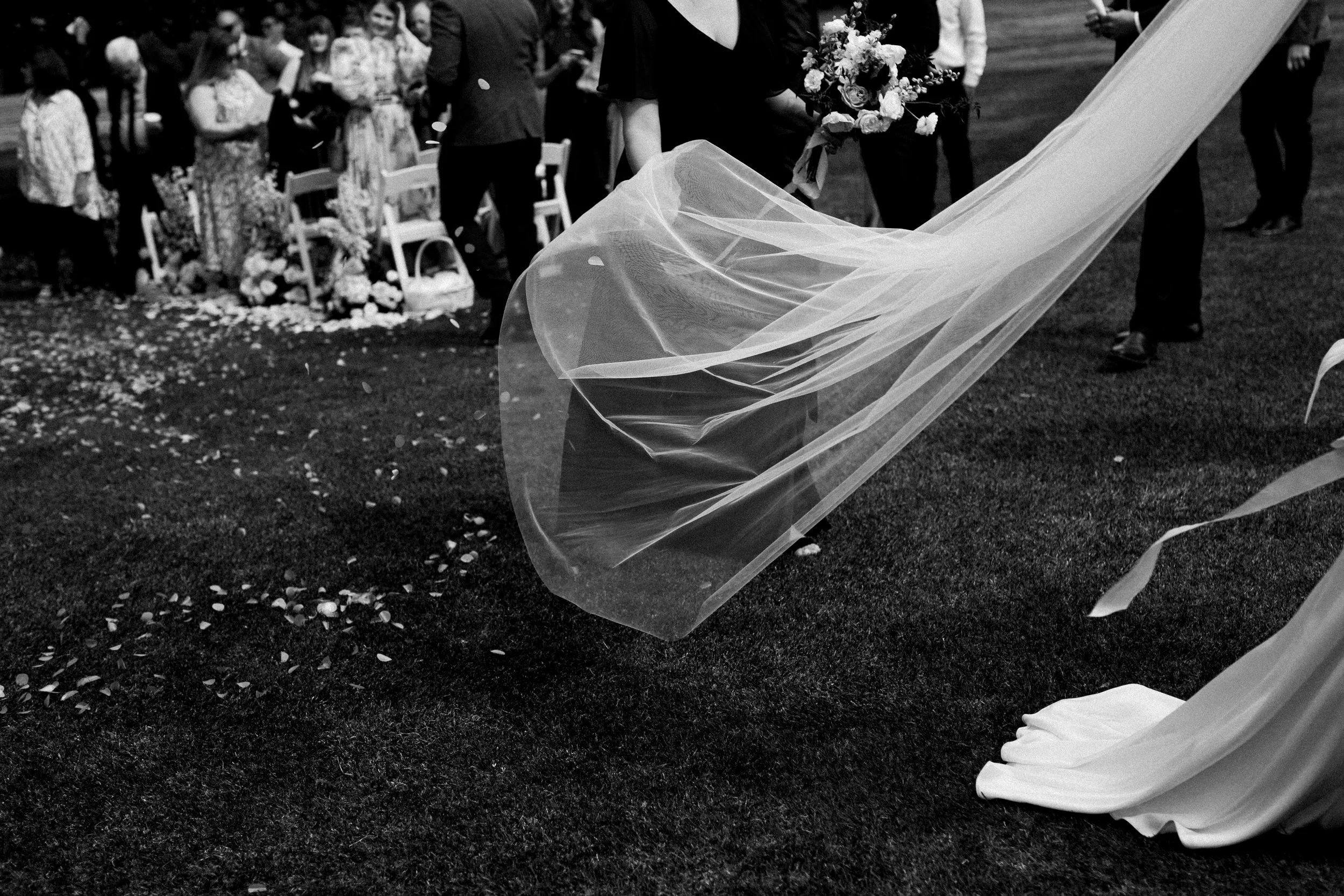 Wedding scene with a bride in a wedding dress and veil, celebrating with guests, some holding bouquets, in an outdoor setting.