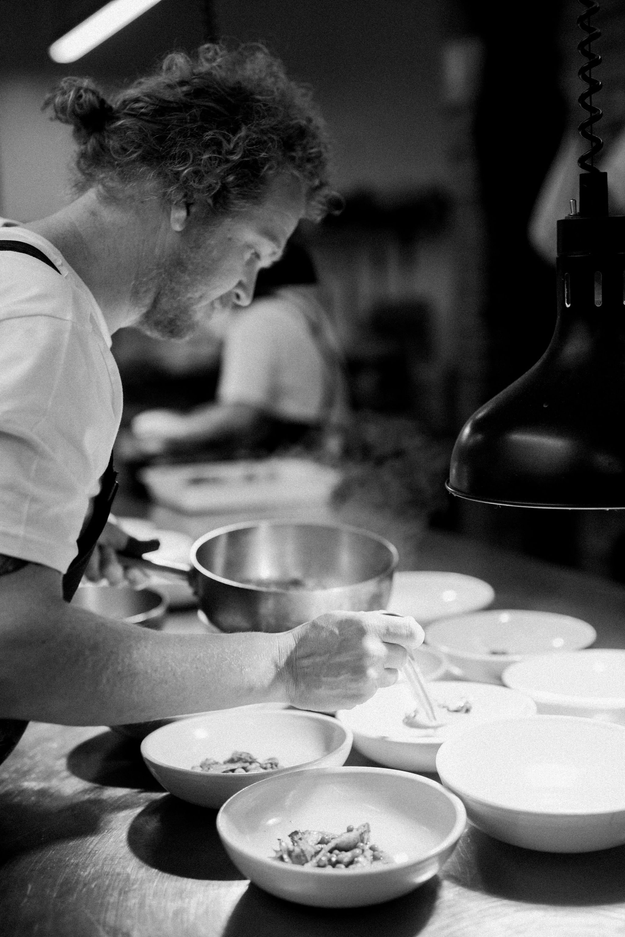 A chef in a kitchen plating a dish at a counter with several emptied bowls in front of him.