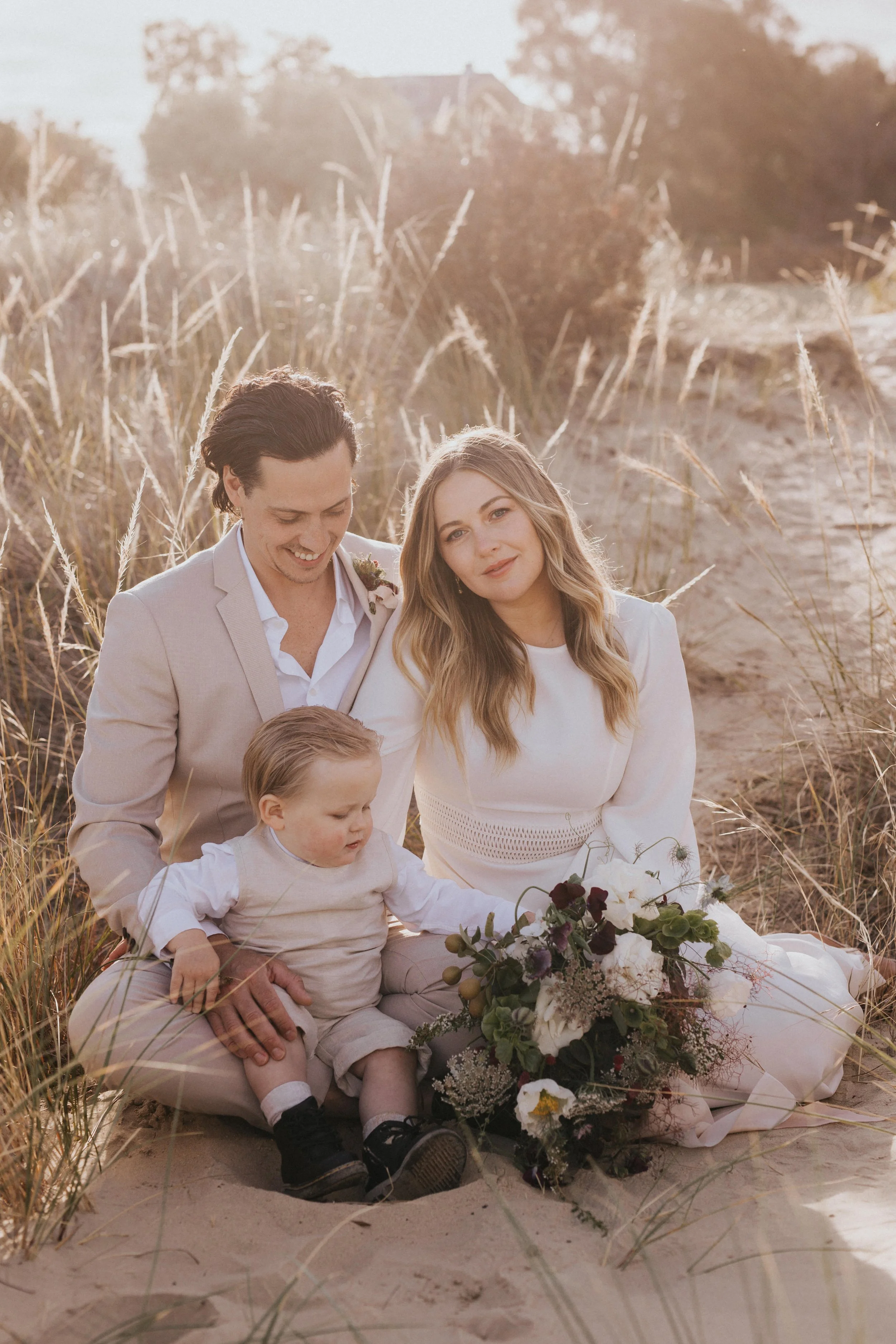 A happy family of three sitting on a sandy beach with tall grasses, celebrating a special occasion with a bouquet of flowers, during golden hour sunlight.