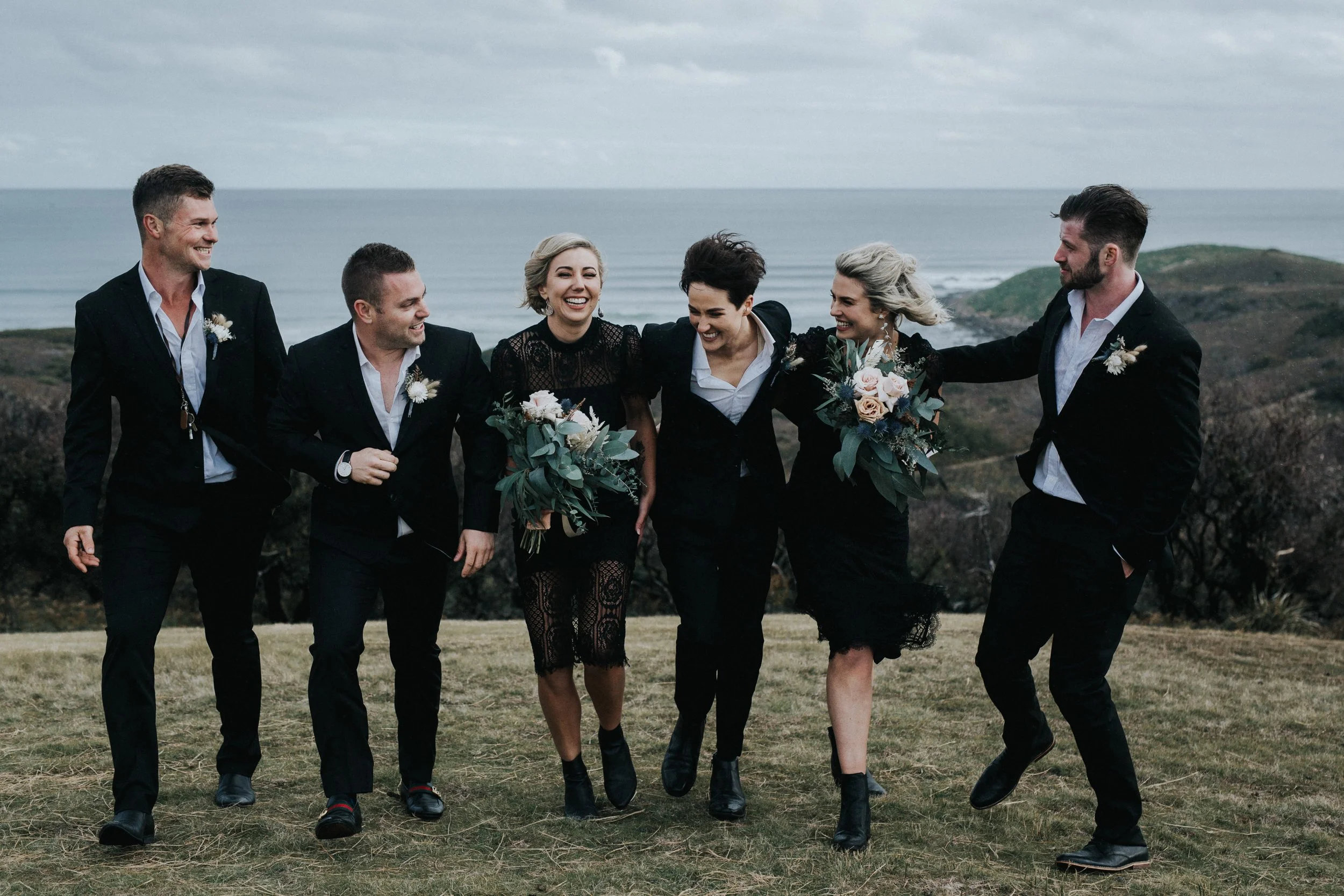 Group of six people dressed in formal black attire, walking and laughing outdoors on a grassy area with the ocean and cloudy sky in the background, holding bouquets of flowers.
