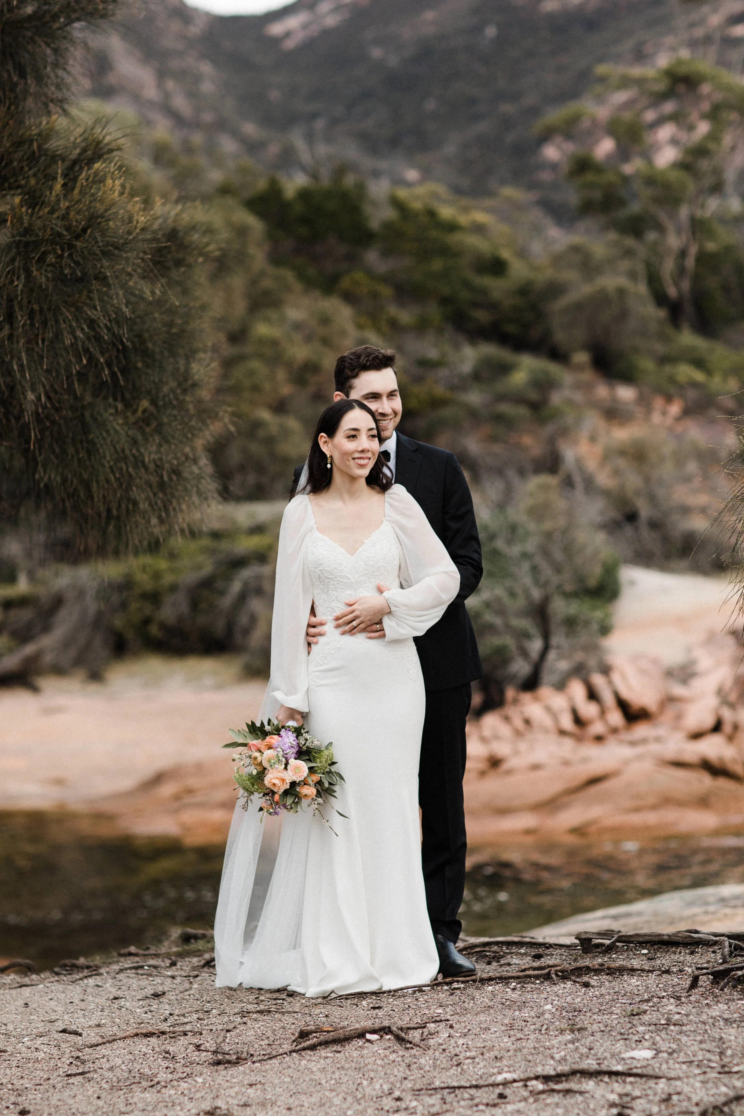 A bride and groom standing outdoors on a rocky terrain with a mountain and trees in the background, smiling after their wedding ceremony.