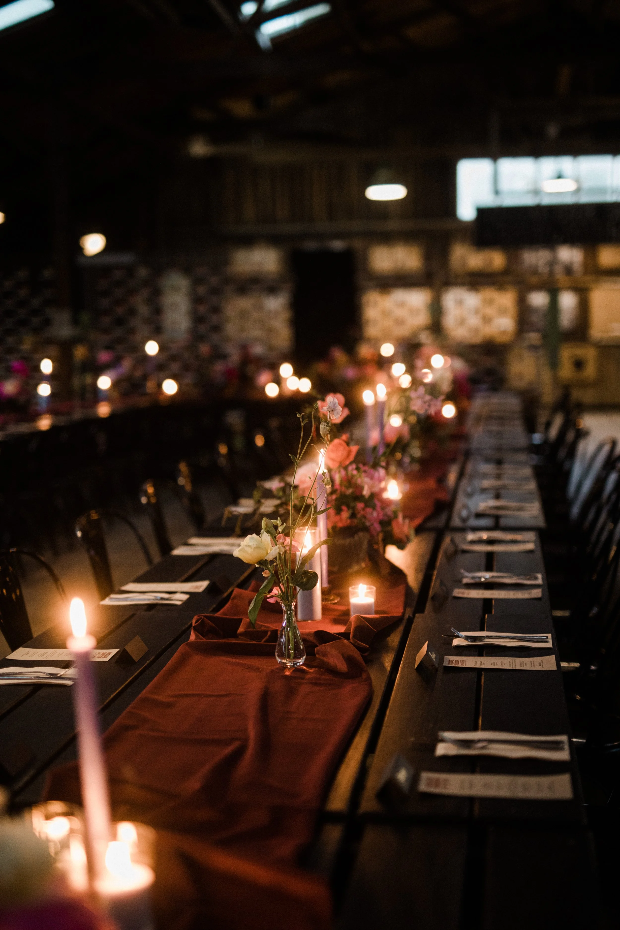 A long dining table set for a formal dinner with floral centerpieces, candles, and place settings in a dimly lit rustic space.