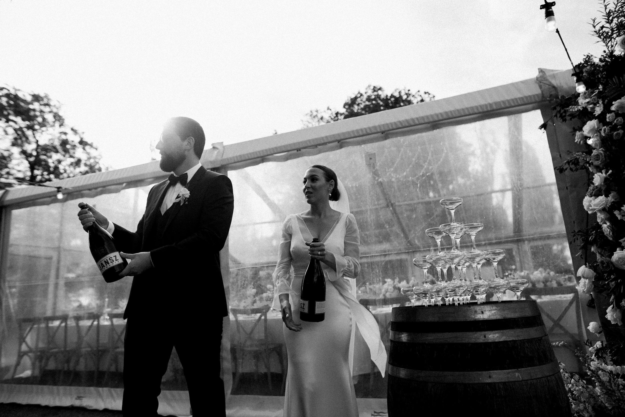 Black and white photo of a newlywed couple at a wedding reception, with the groom opening a champagne bottle and the bride holding another bottle; champagne glasses are stacked on a barrel nearby, with floral decorations and string lights in the back