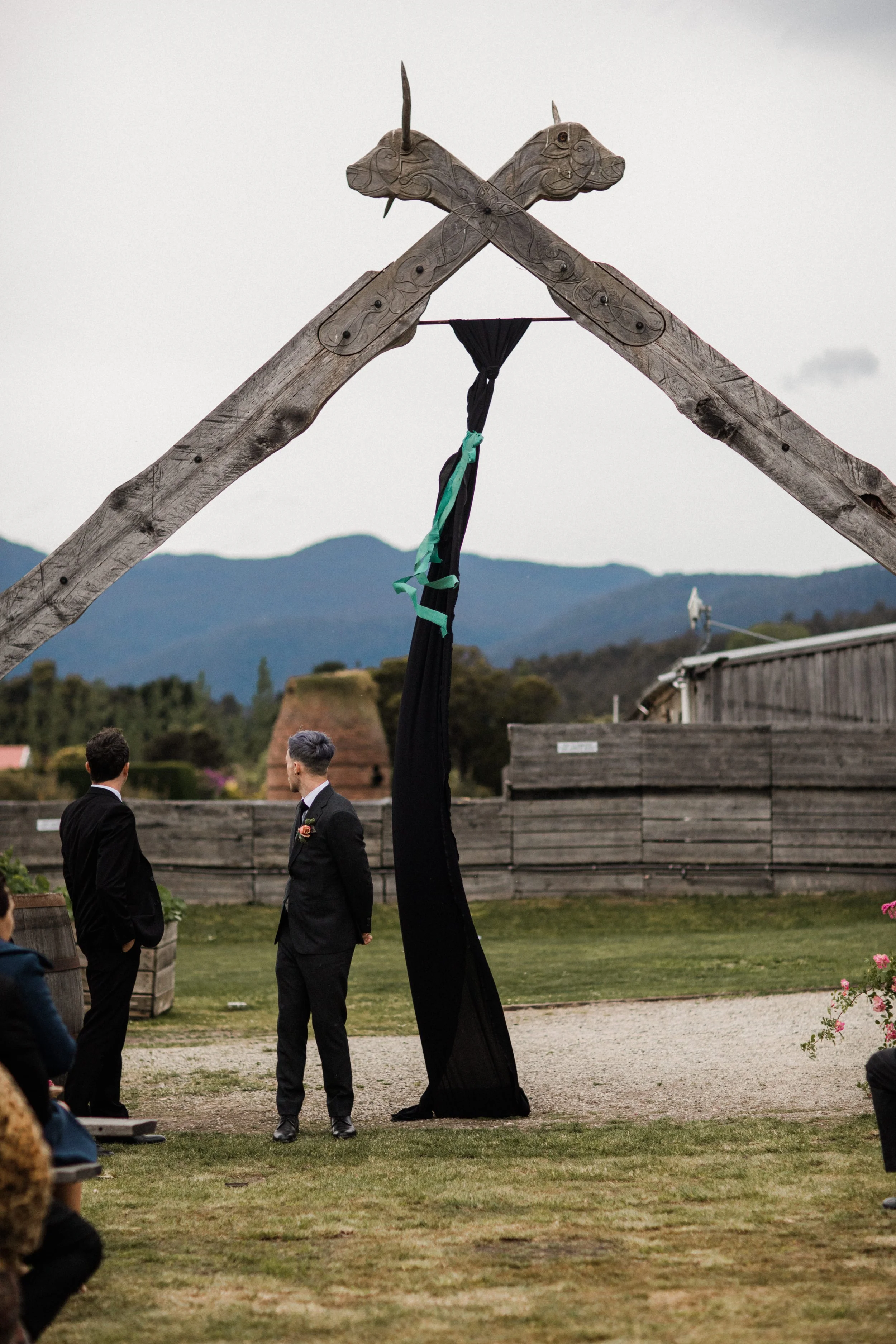 Two men in black suits and a third partially visible person are standing outside near a wedding altar made of wooden beams with carved unicorn heads, with a mountain range in the background.