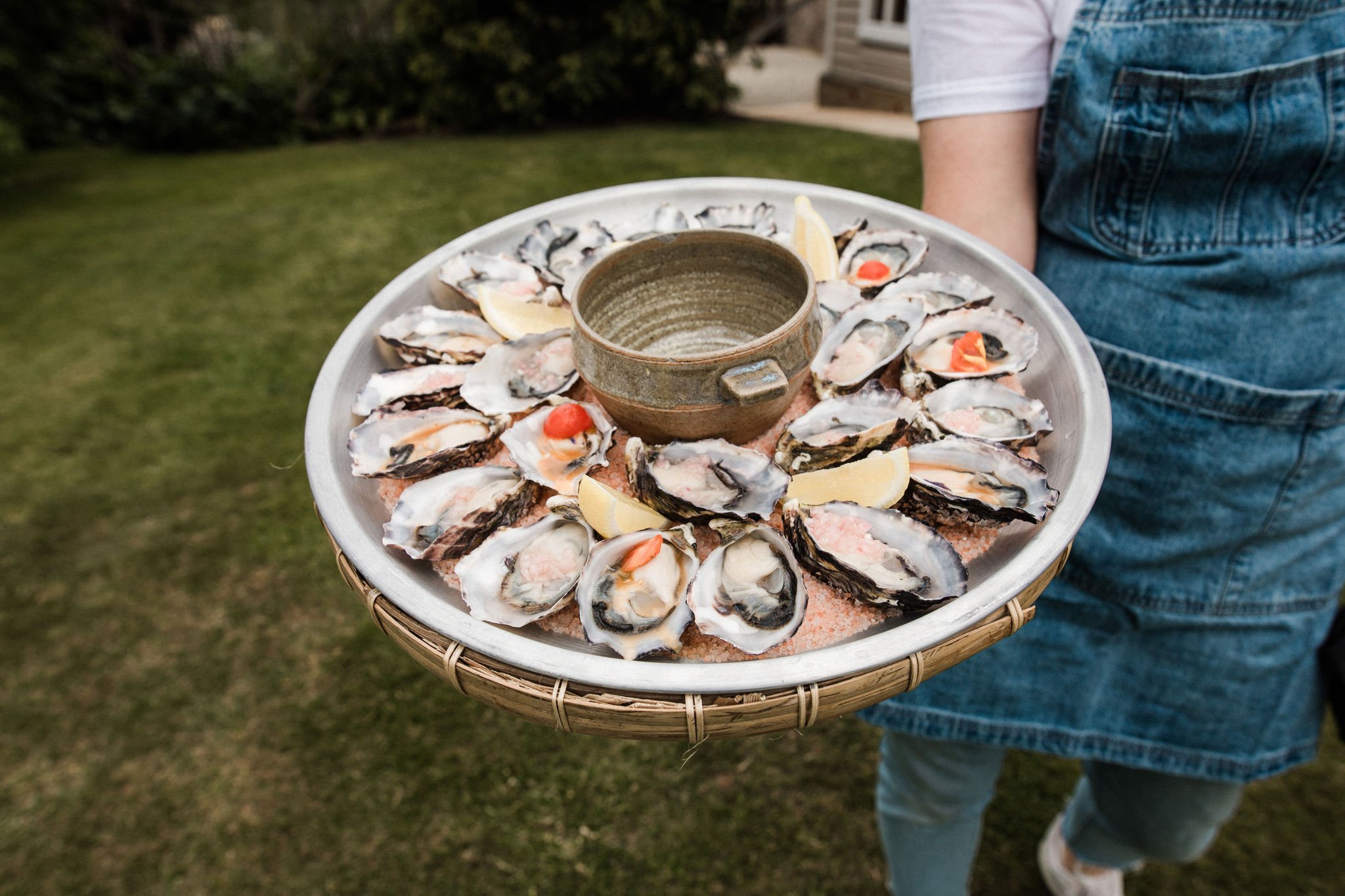 Person holding a large tray of fresh oysters garnished with lemon wedges and small red toppings, with a small ceramic bowl in the center on an outdoor grassy area.