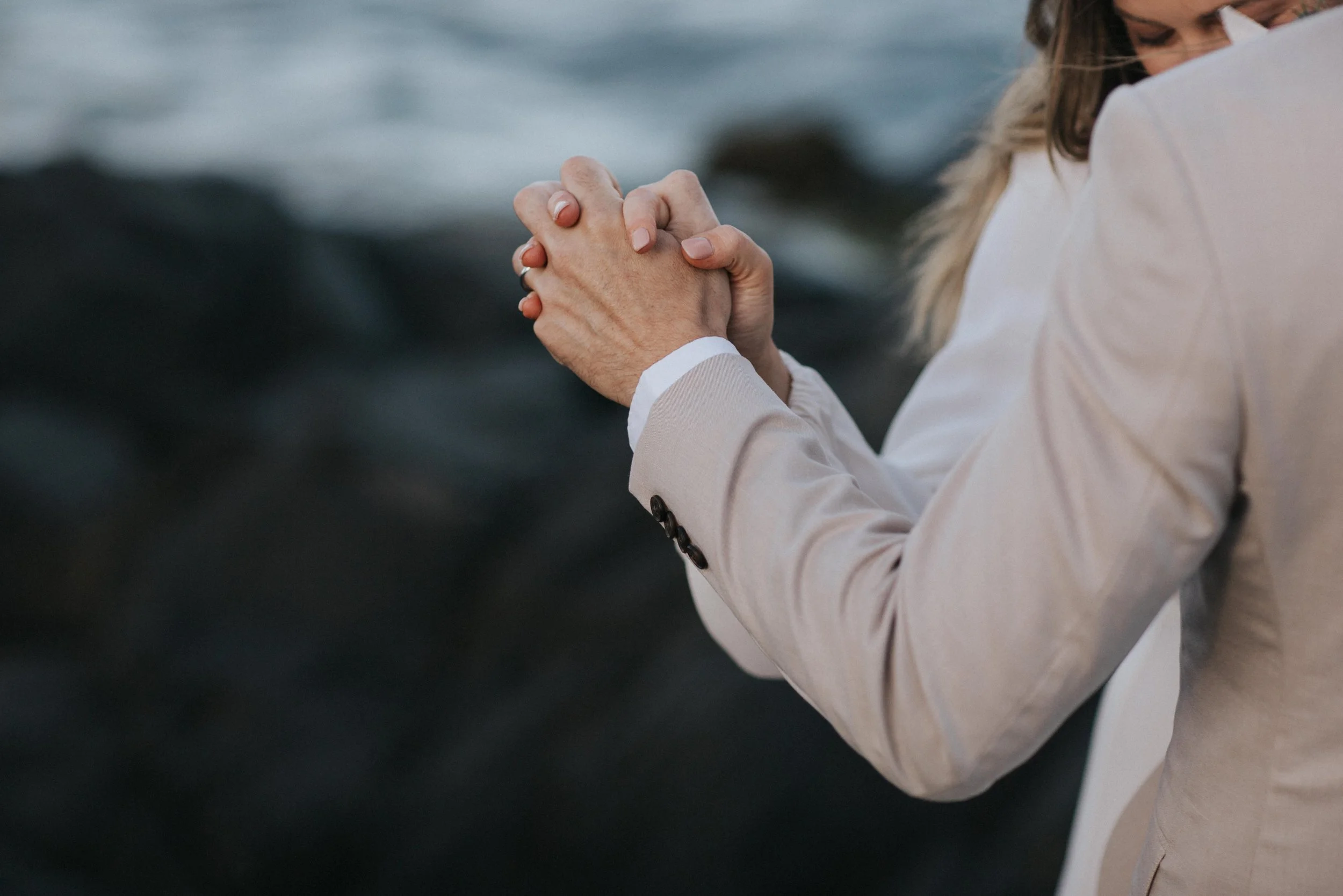 A person in a beige suit with their hands clasped together during prayer or reflection, with a blurred background of water.