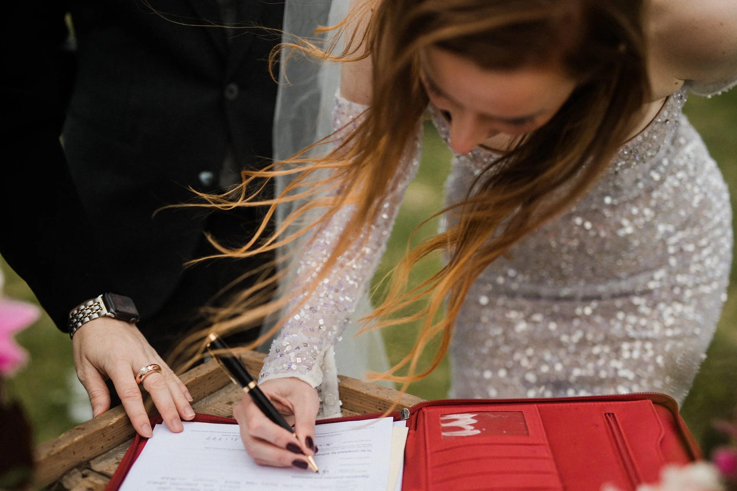 A woman with long red hair in a wedding dress signs a document on a wooden table, with a person beside her helping or observing.