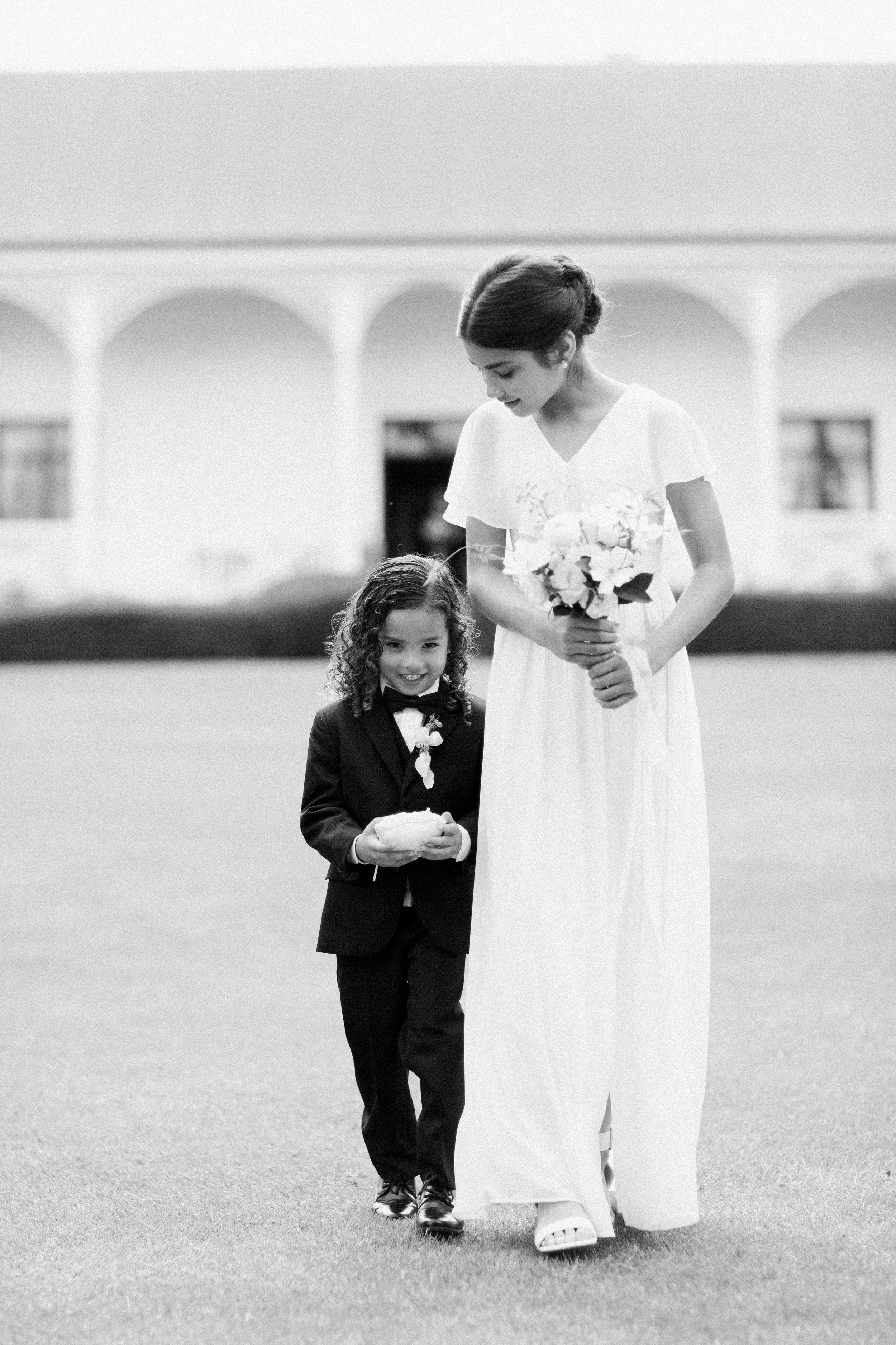 A woman in a white dress holding a bouquet and a young boy in a suit walking outdoors during a wedding ceremony.