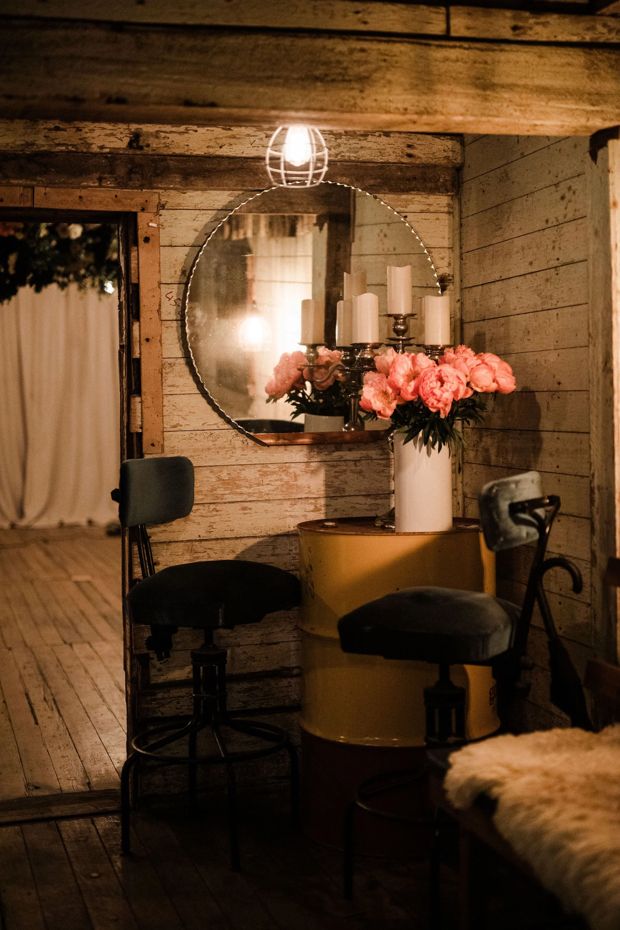 A rustic interior featuring a round mirror, pink peony flowers in a tall white vase, and a candelabra with white candles on a yellow barrel table. There are two black chairs and a furry cushion, with warm ambient lighting from a ceiling bulb and refl