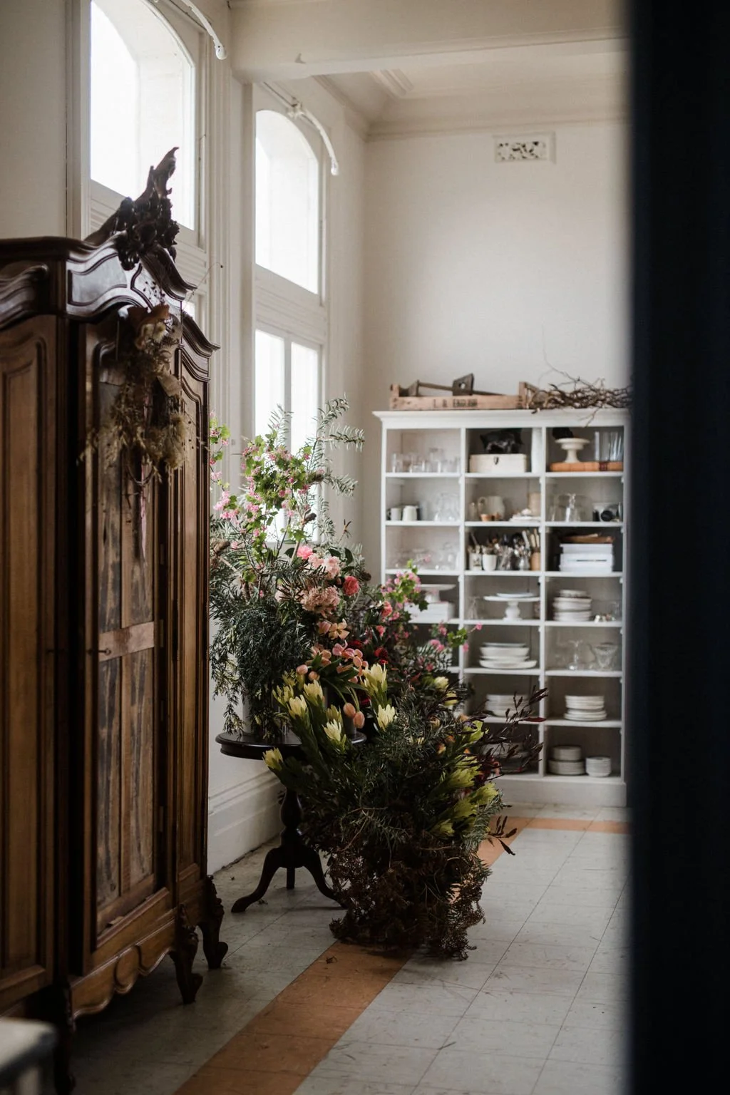 A corner of a room with large windows, an ornate wooden wardrobe, a white shelving unit filled with dishes and glassware, and a large floral arrangement on the floor.