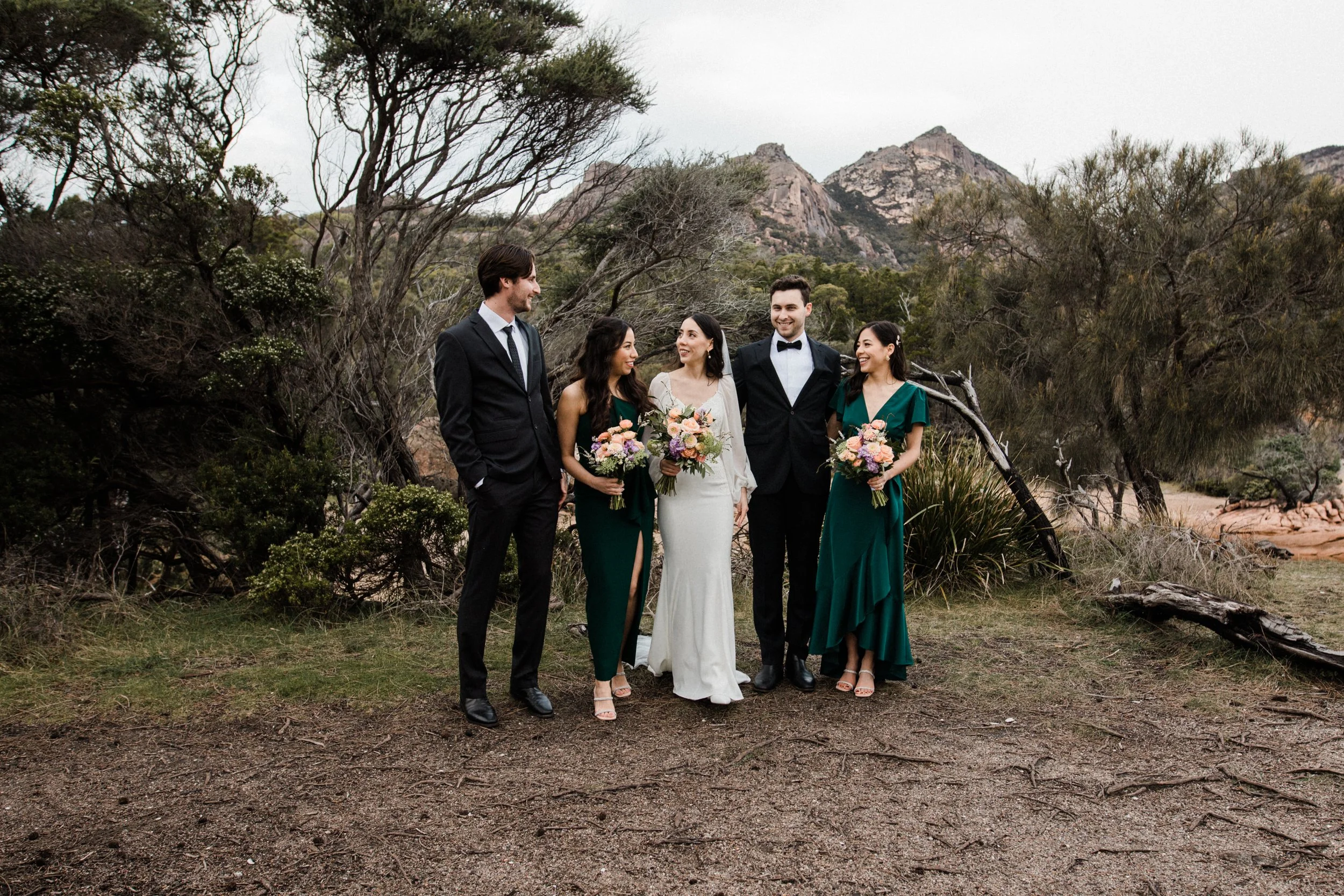 A wedding party of six people standing outdoors on dirt ground surrounded by trees and mountains, with three women holding bouquets and two men in suits, and one woman in a white wedding dress.