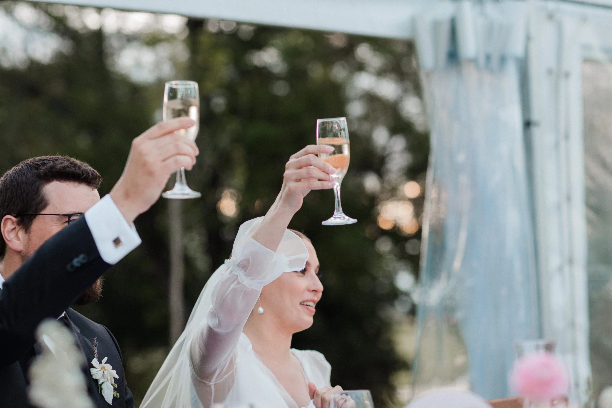 A bride and groom raising champagne flutes during a wedding toast outdoors, with a decorated table and greenery in the background.
