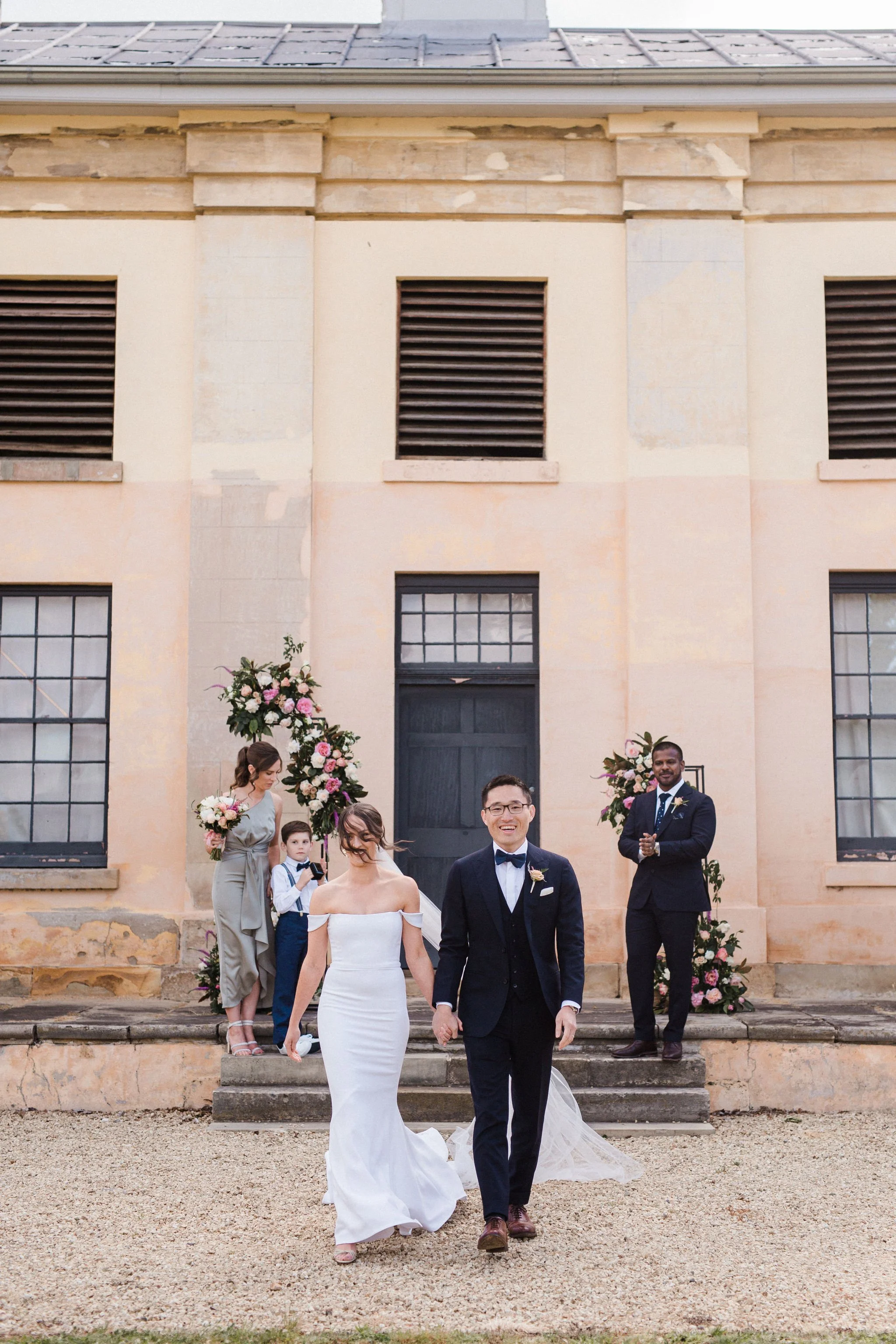 Bride and groom walking hand in hand in front of an old building during their wedding, with wedding party members and floral decorations in the background.