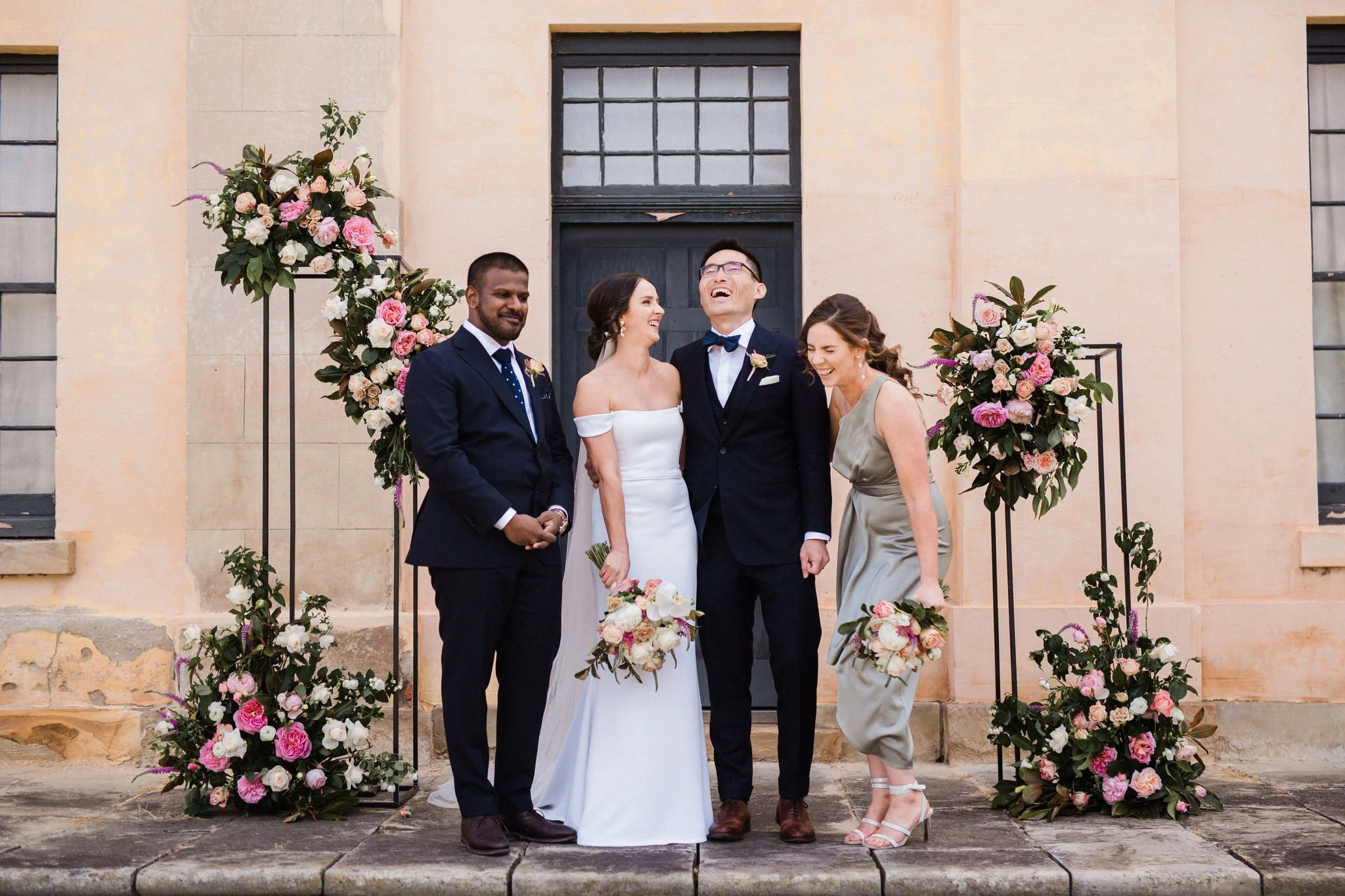 A wedding group standing in front of a building decorated with floral arrangements, including a bride in a white off-shoulder gown holding a bouquet, a groom in a black tuxedo, and two guests, a man in a navy suit and a woman in a silver dress, all s