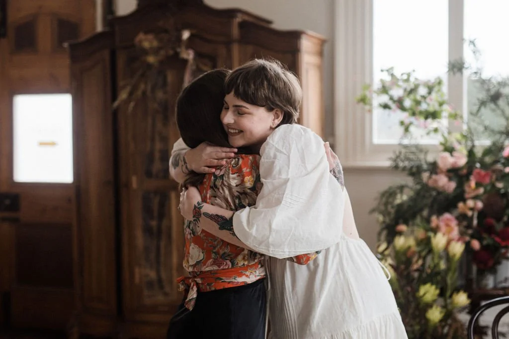 Two women hugging and smiling in a room with wooden furniture and large floral arrangements.