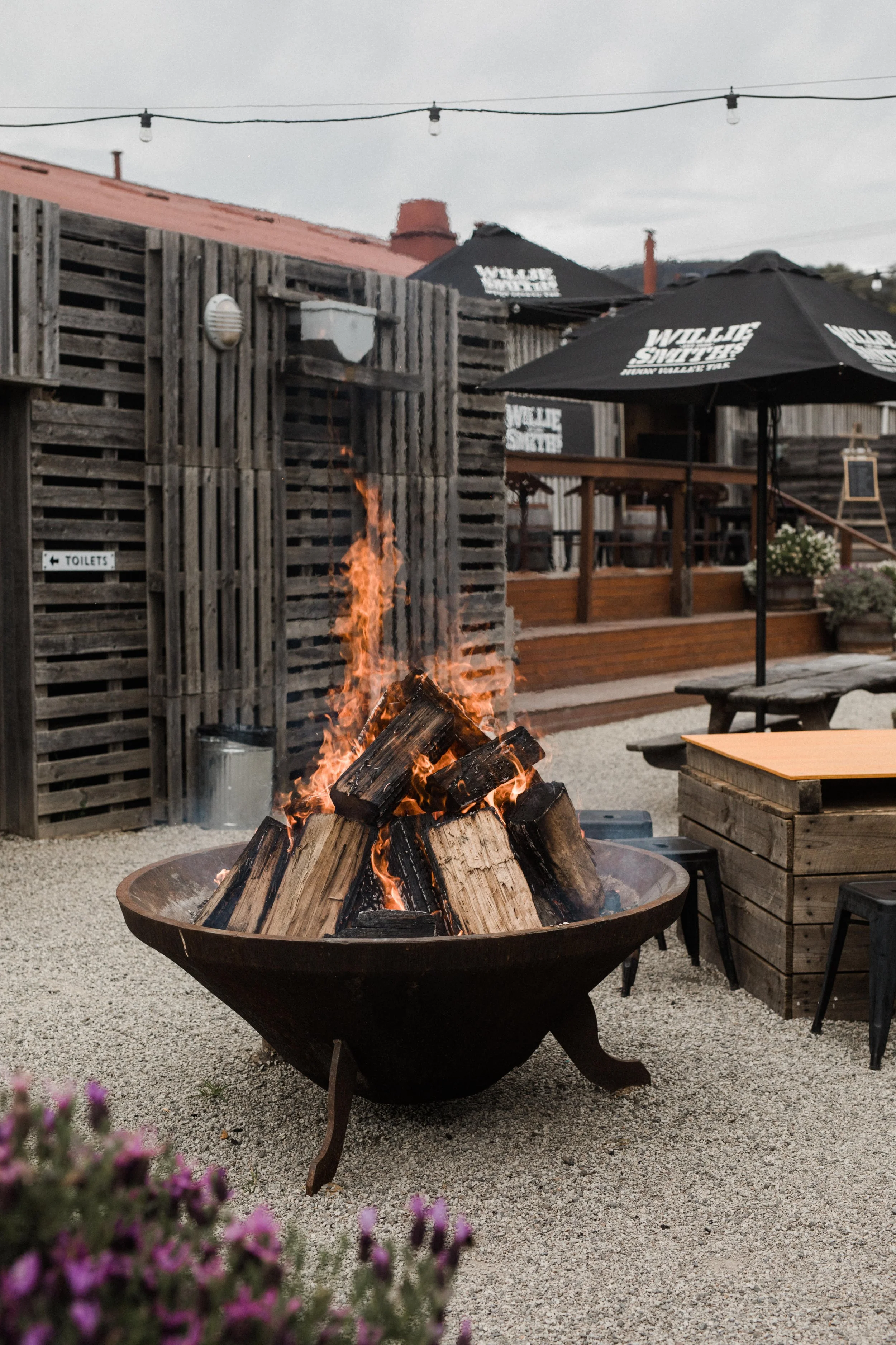 An outdoor patio area with a fire pit filled with burning logs, surrounded by wooden tables and chairs, umbrellas for shade, and string lights overhead at dusk or evening.