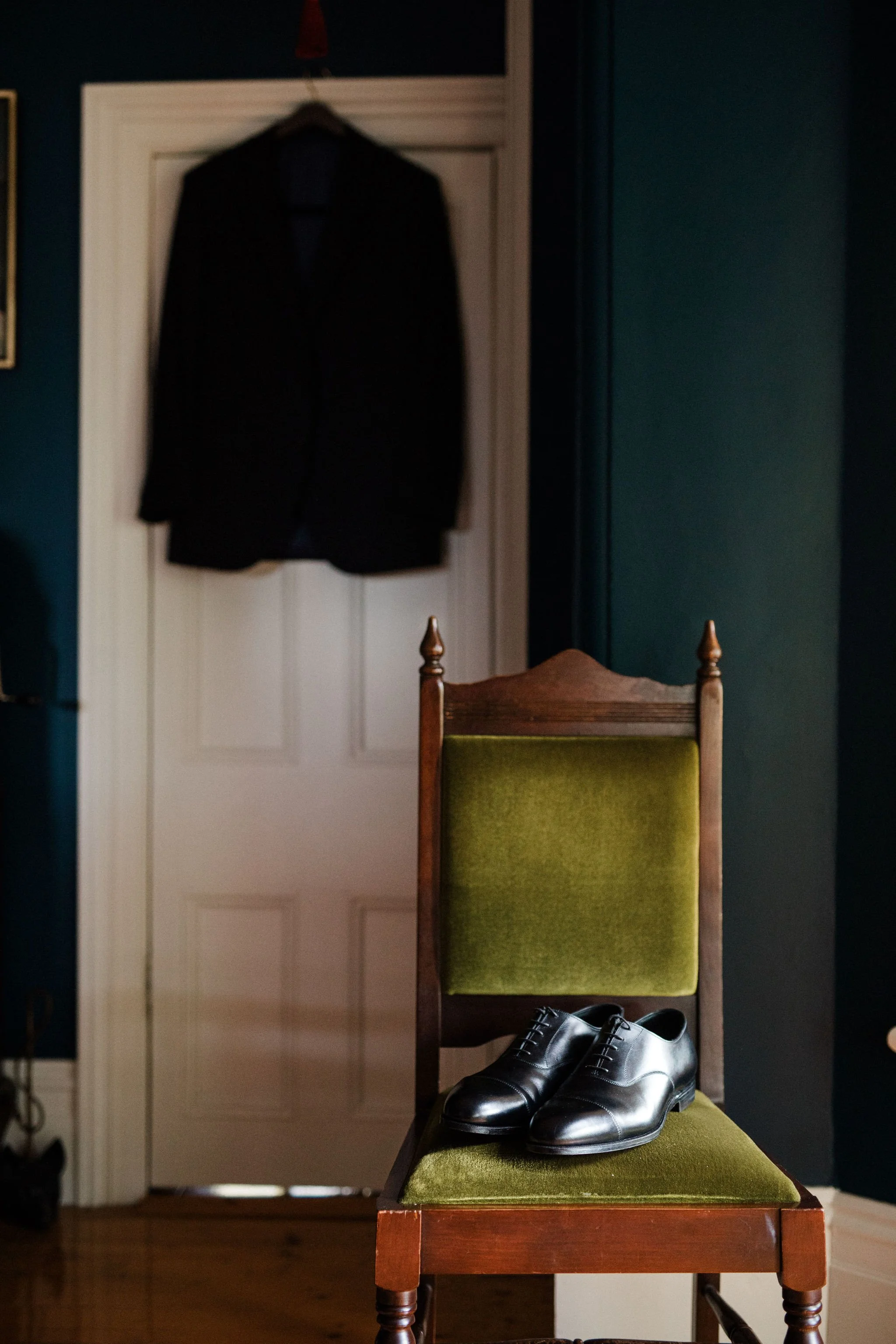 A pair of polished black dress shoes placed on a green upholstered wooden chair, with a black blazer hanging on the door in the background.