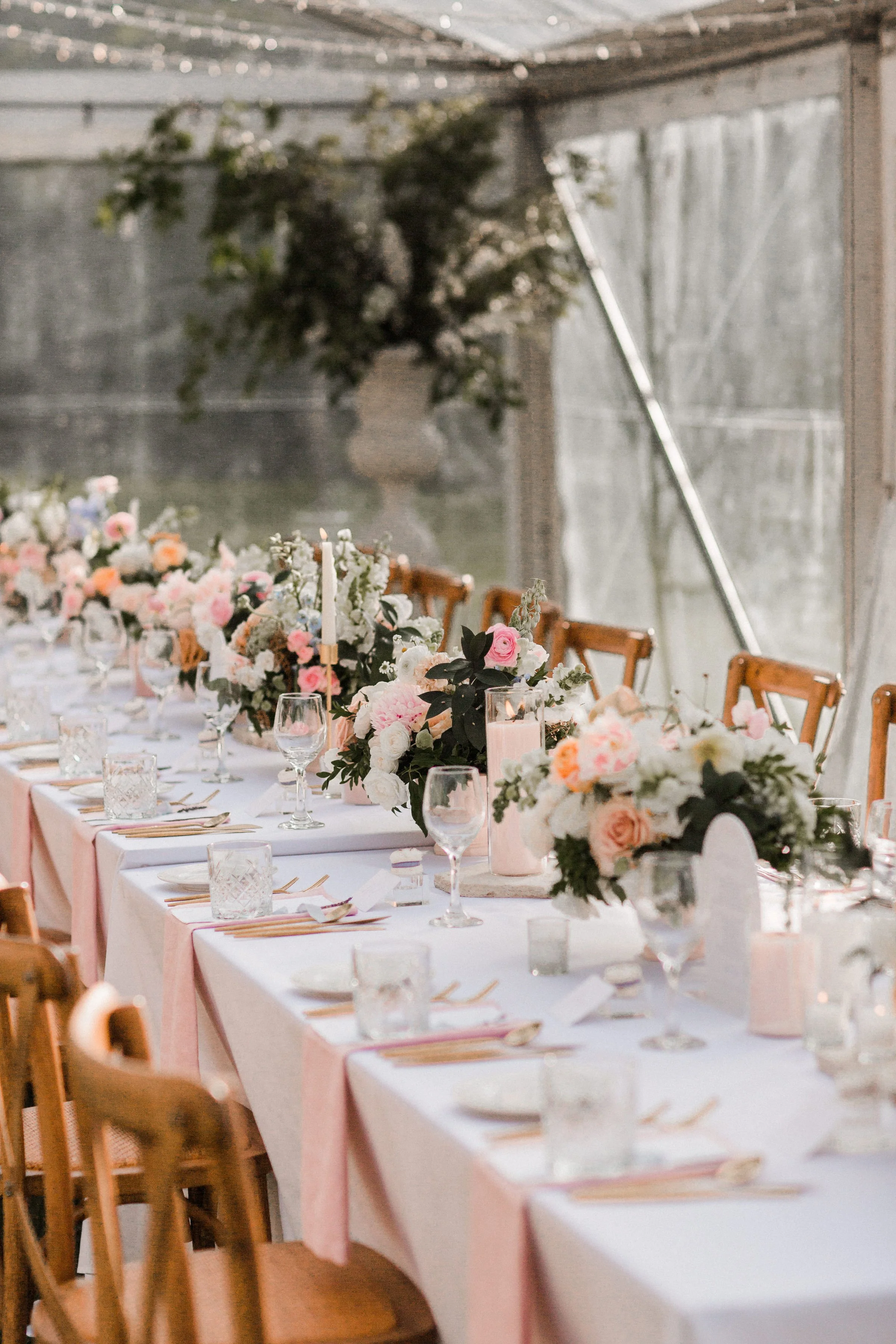 Long dining table decorated with pink and white flowers, candles, and glassware inside a rustic tent.