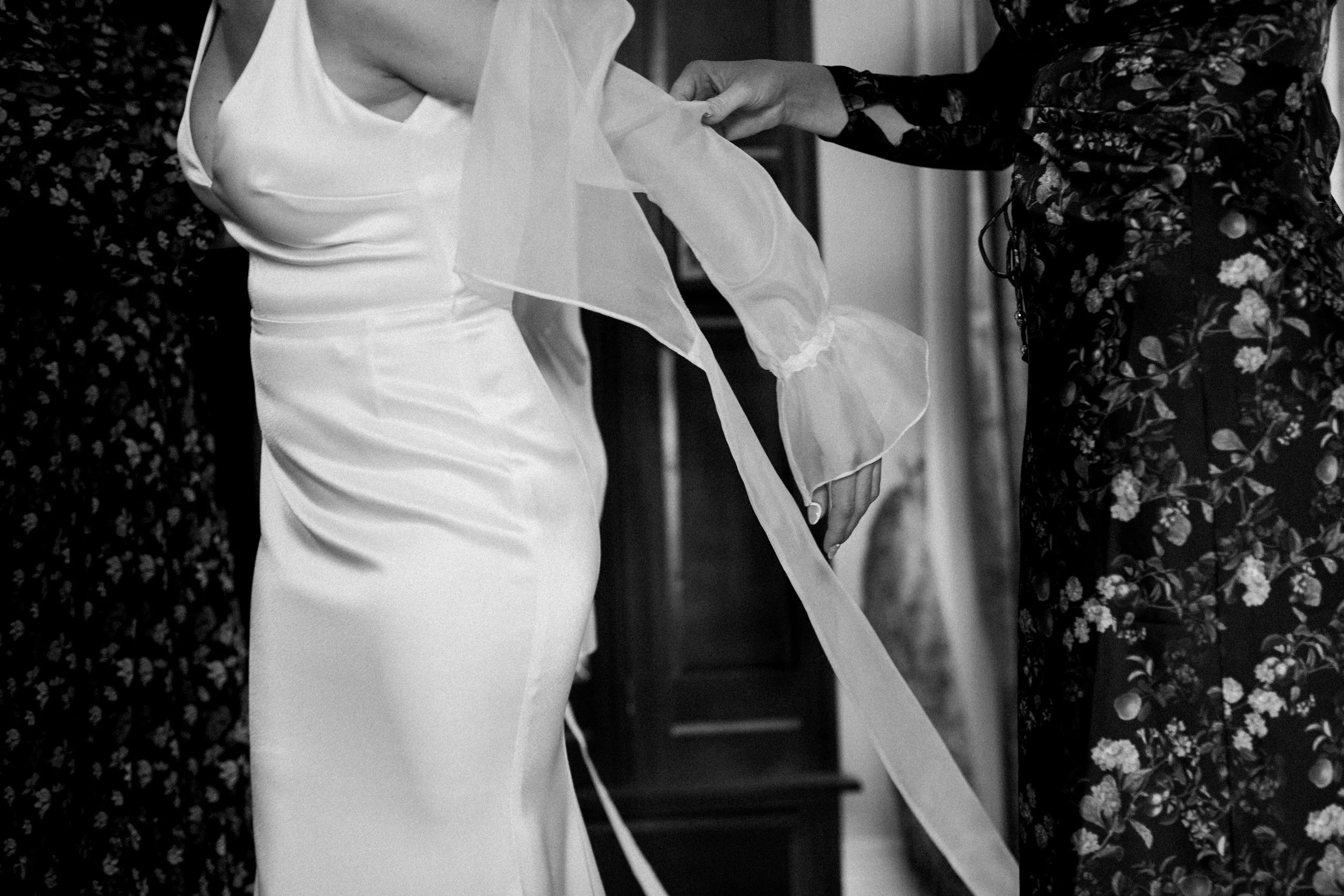 Close-up of a bride in a satin wedding dress holding hands with another person wearing a floral dress, possibly during a wedding ceremony.