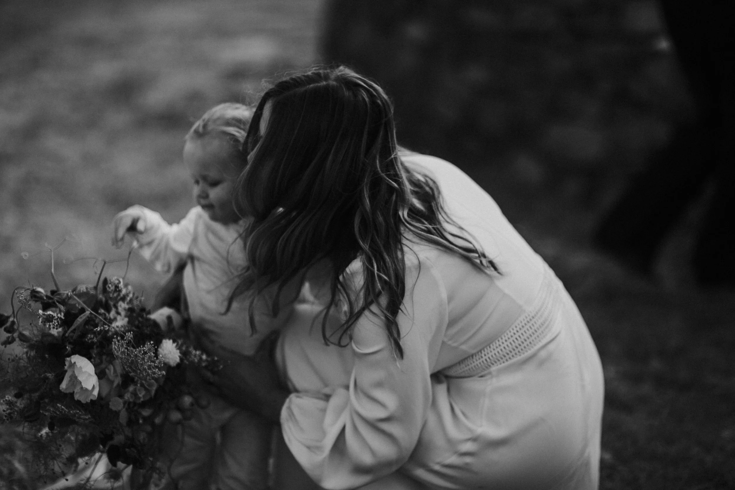 A woman with wavy hair bending down to assist a young girl in picking flowers, surrounded by grass and blurred background.