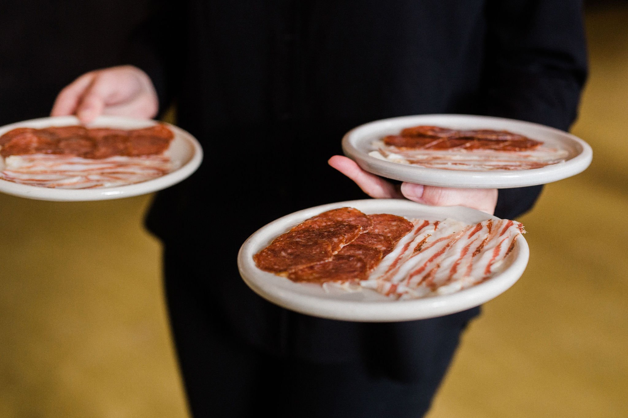 Person holding three small white plates with slices of cured meat, such as bacon and salami, against a blurred yellow background.