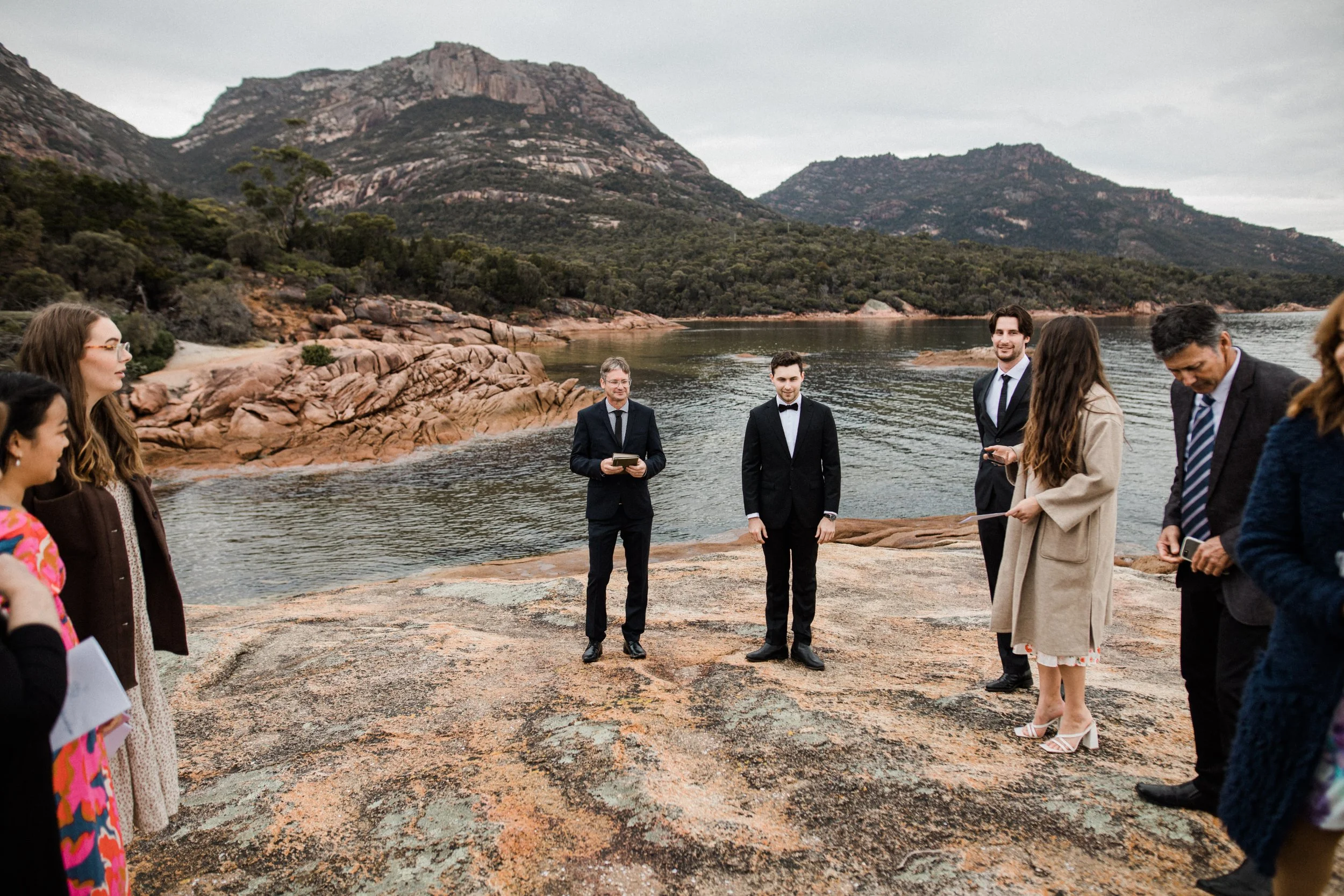 A wedding ceremony taking place outdoors on a rocky shoreline with a lake and mountains in the background. The bride and groom stand at the center, with the officiant and guests surrounding them.