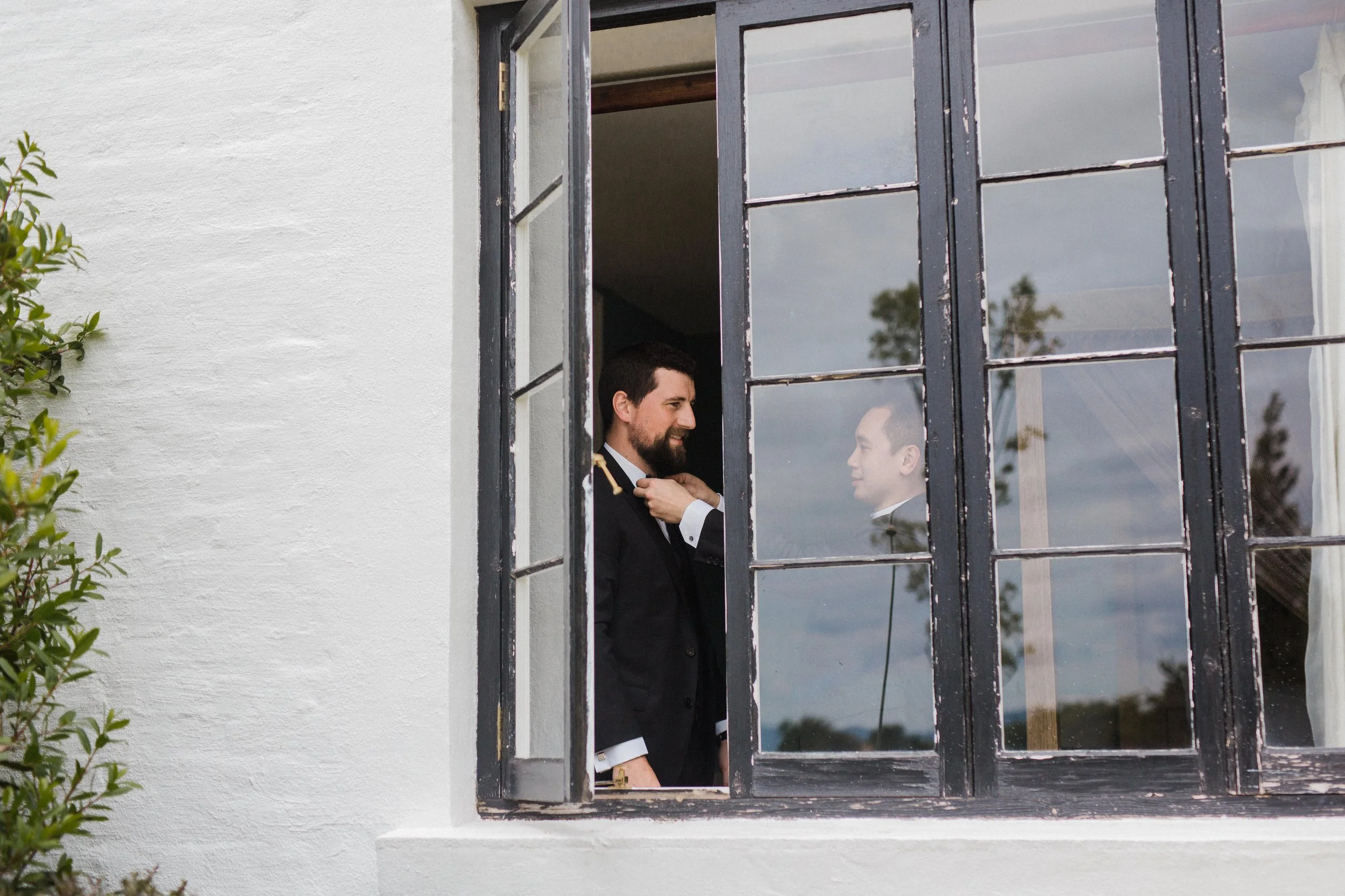 Two men in suits, one helping the other adjust a bow tie through a window.
