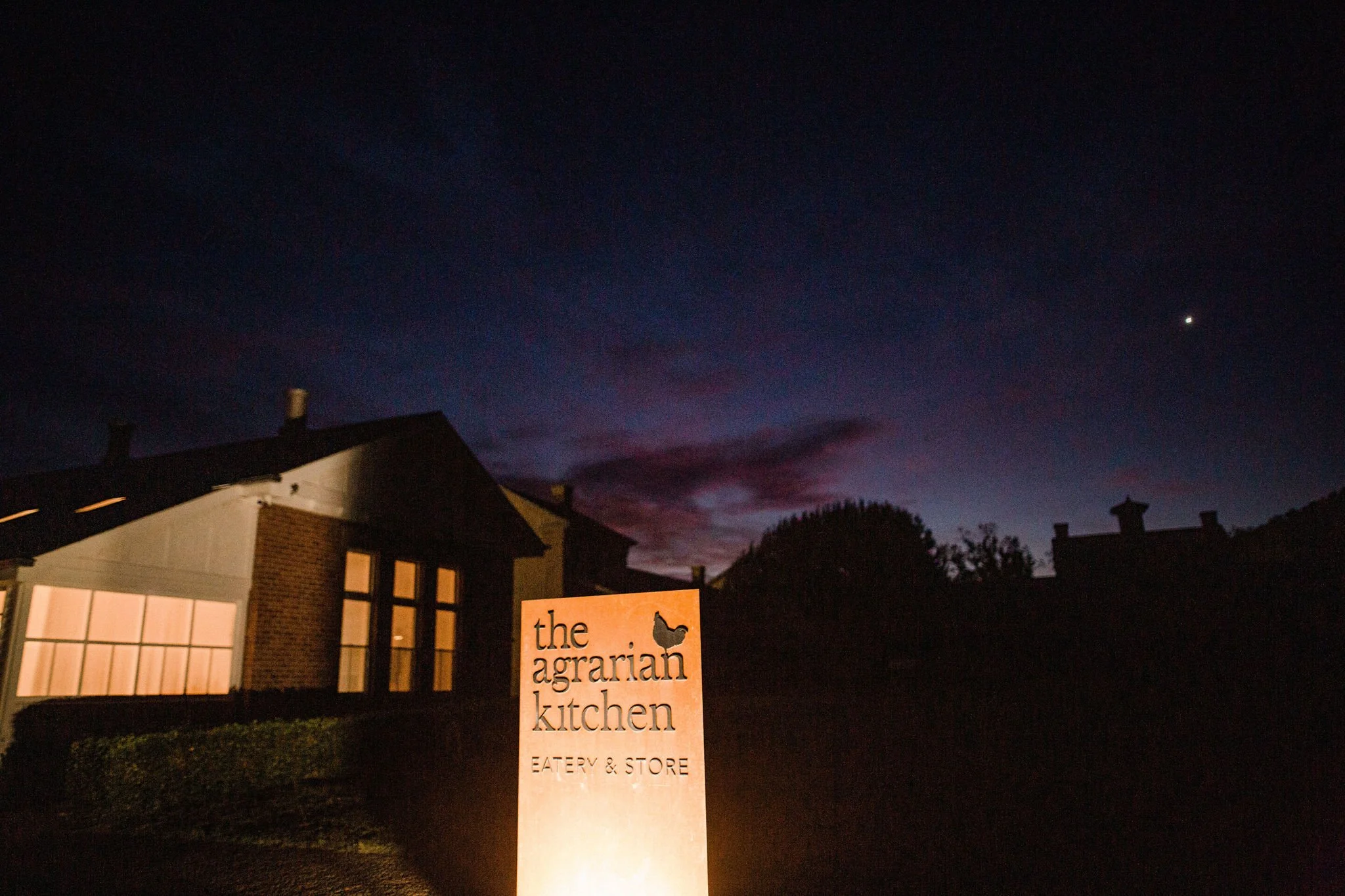A sign for 'the agrarian kitchen Eatery & Store' in front of a building at dusk, with a dark blue and purple sky and a bright planet or star visible.