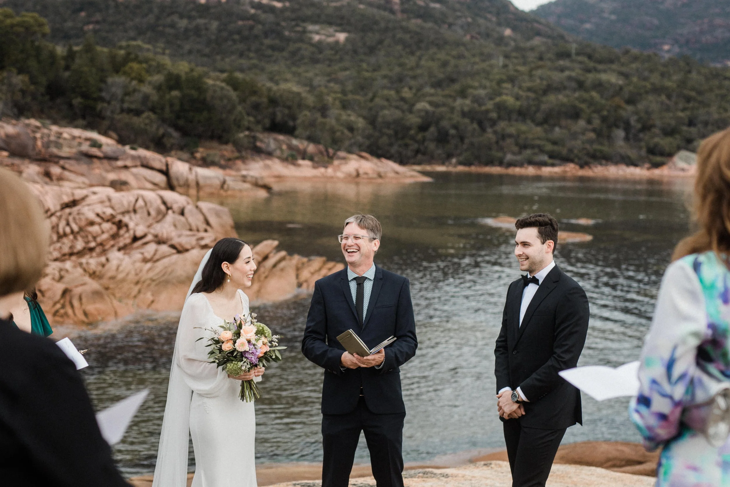 A wedding ceremony taking place outdoors by a lake with rocky shores and mountainous background, featuring a bride in a white dress holding a bouquet, a groom in a black tuxedo, and an officiant in a suit.