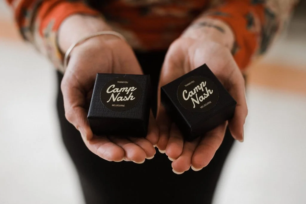 Person holding two small black boxes labeled "Thank You Camp Nash Melbourne"
