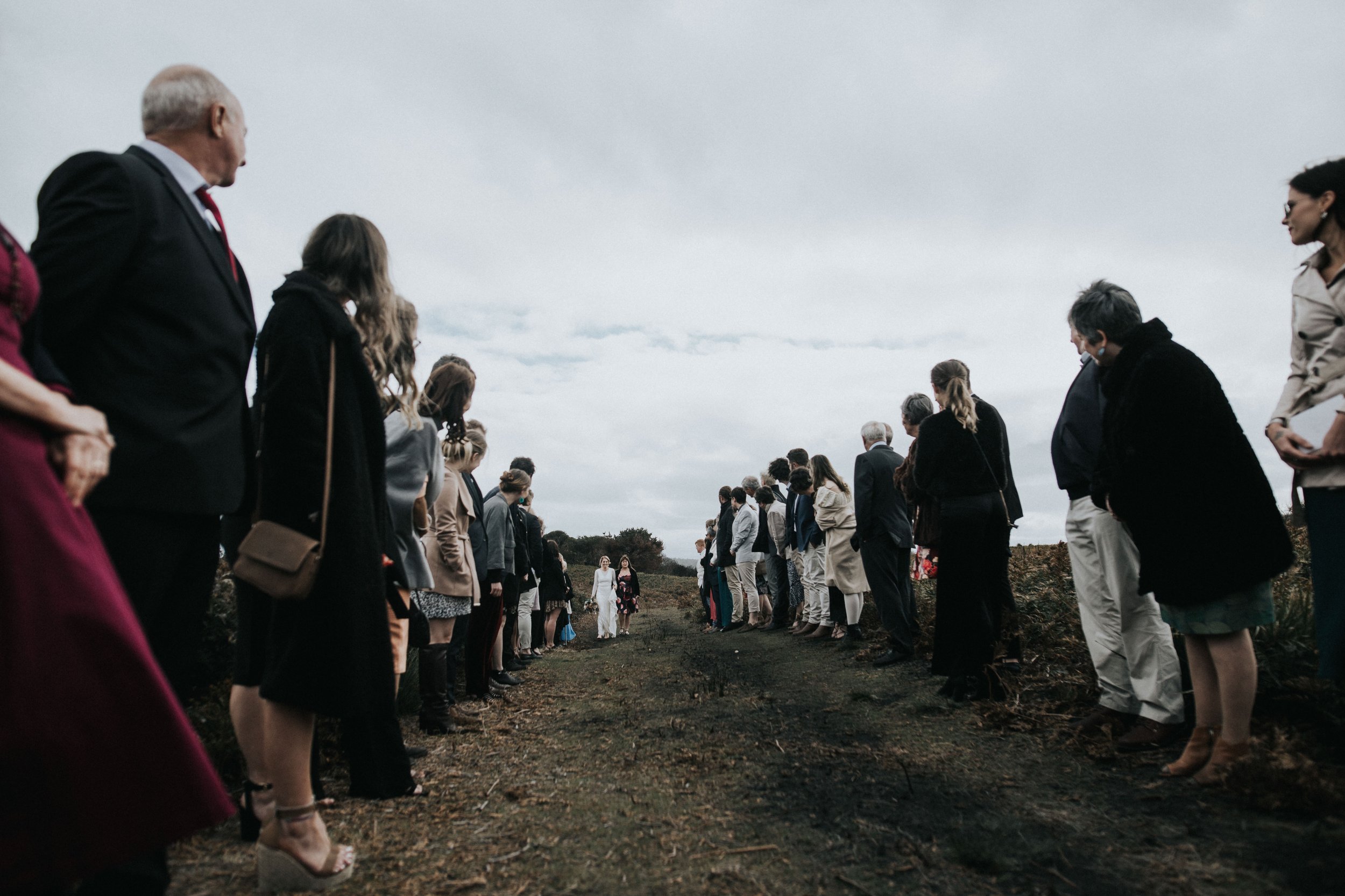 A large group of people dressed in formal attire standing on either side of a dirt path outdoors, forming a corridor with couple walking in the distance.