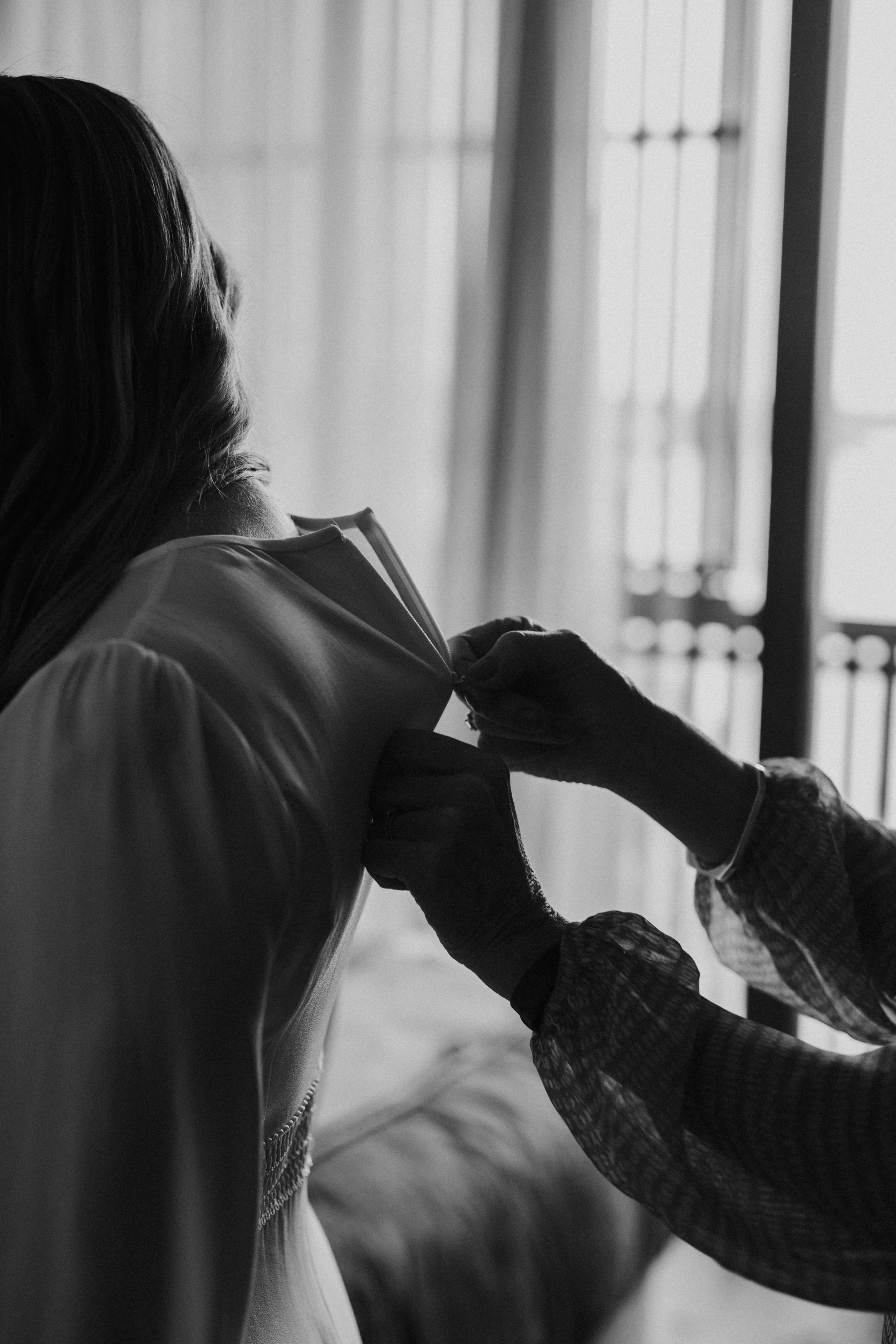 Person helping another put on a wedding dress as sunlight streams through window blinds.