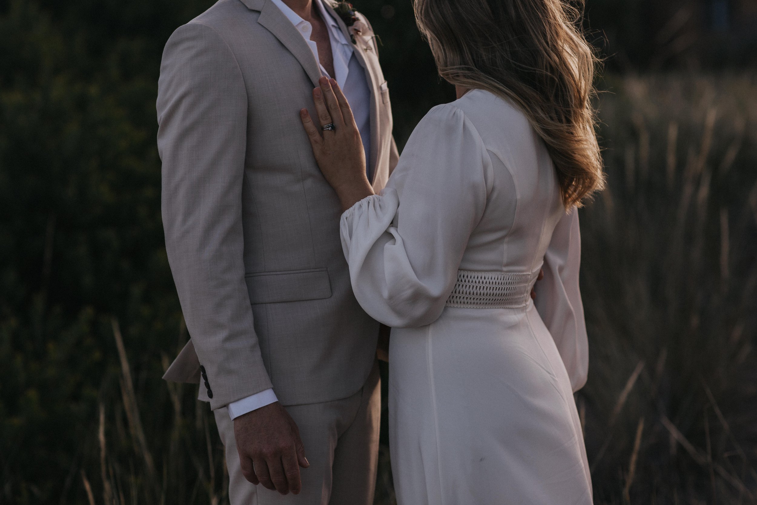 A couple dressed in white, standing close with the woman touching the man's chest. The scene appears to be outdoors during dusk or early evening.