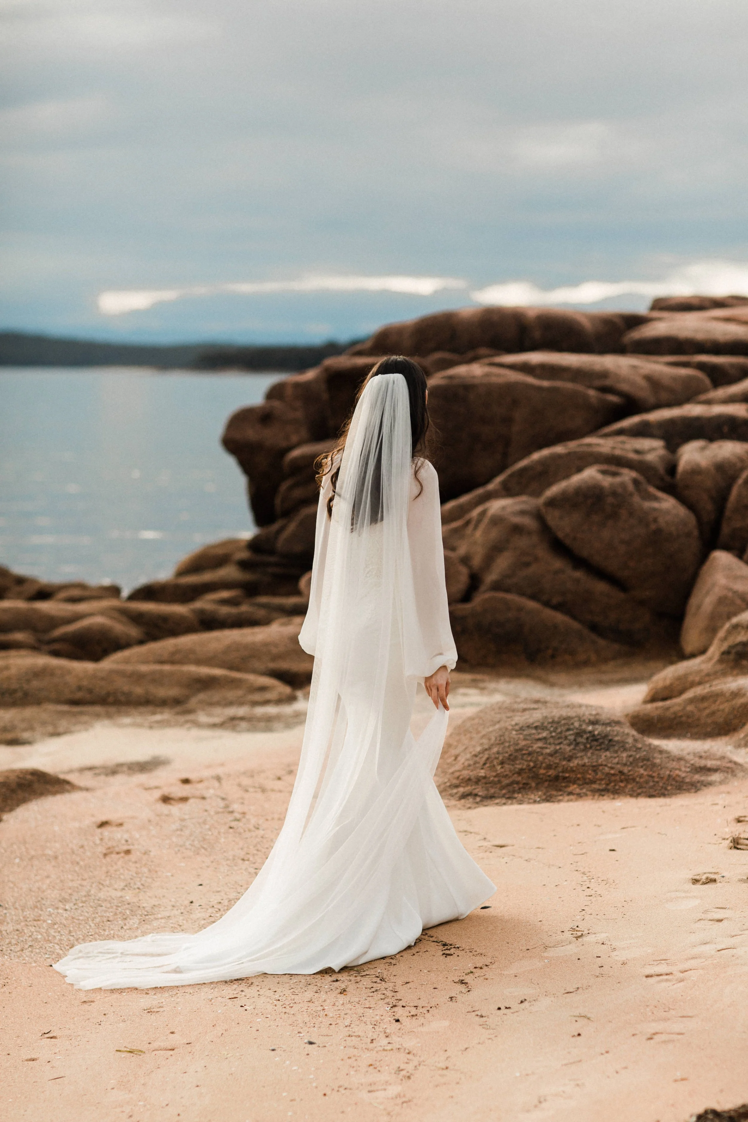 A woman in a white wedding dress with a veil standing on a sandy beach near large rocks, looking out at the water and cloudy sky.