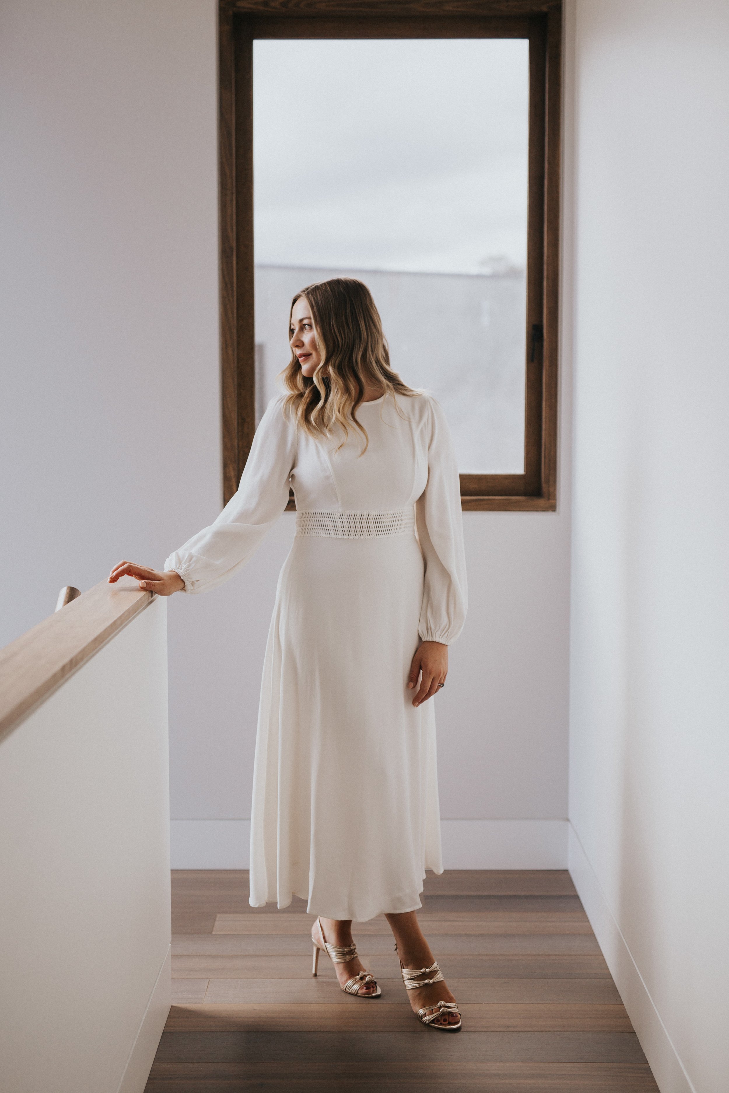 A woman in a long white dress standing at a window, smiling and looking to her left, in a minimalist indoor setting.