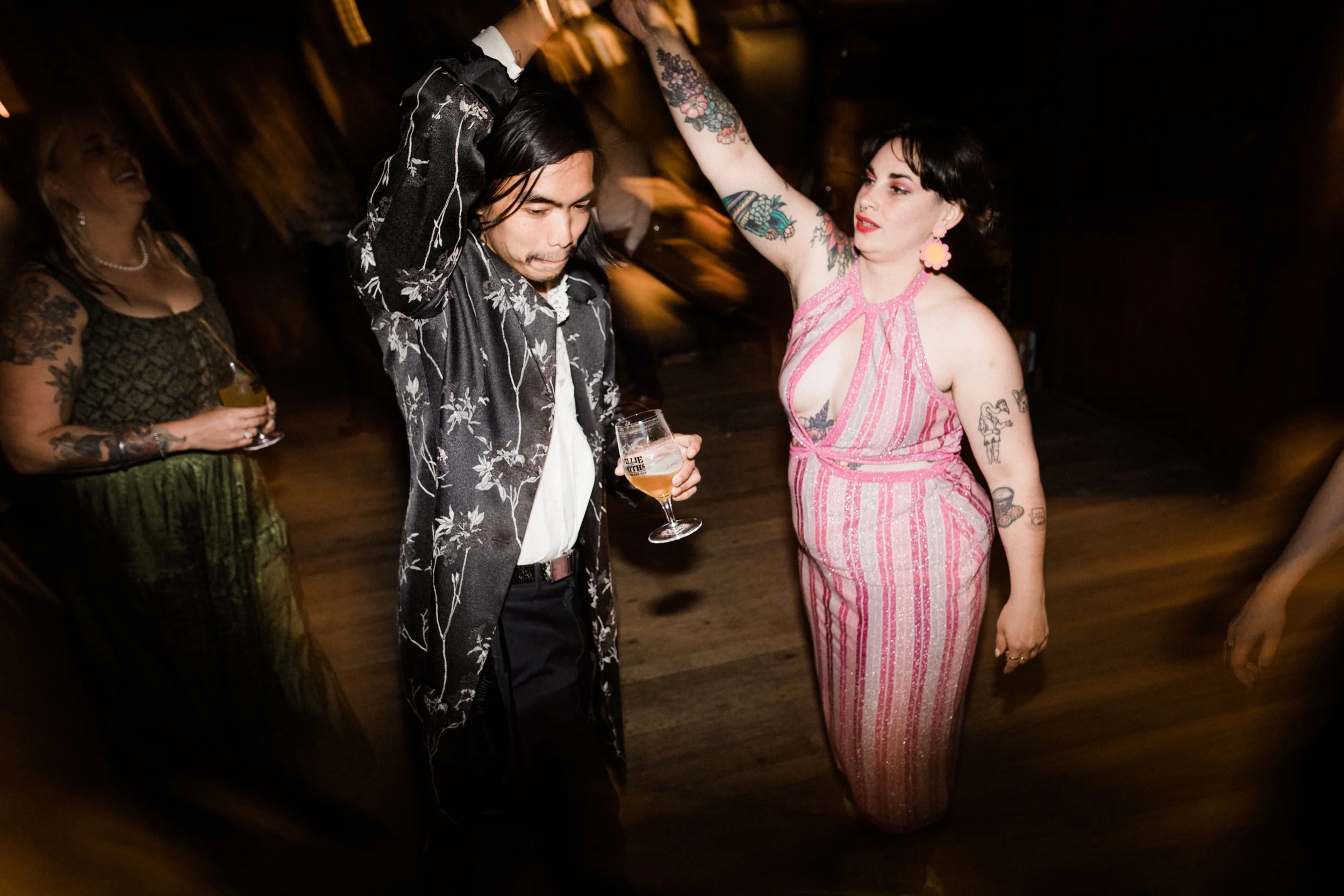 A man and woman dancing together at a party. The woman is wearing a pink and white striped dress with cutouts, and the man is wearing a black floral jacket. The woman has dark hair and tattoos on her arms, while the man has long hair and is holding a