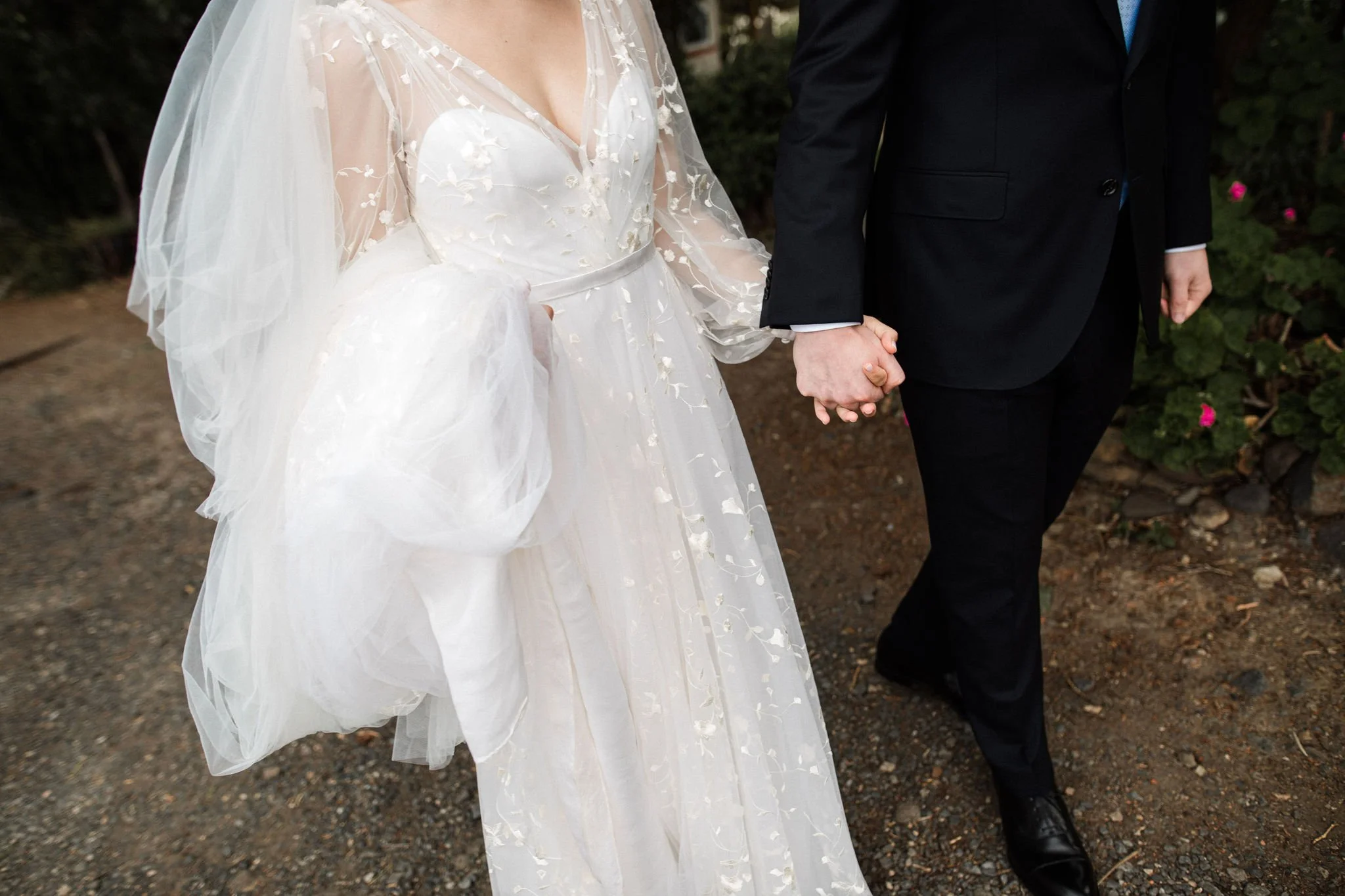 A bride and groom holding hands during a wedding ceremony, with the bride wearing a white wedding dress with sheer, embroidered sleeves and the groom in a black suit.
