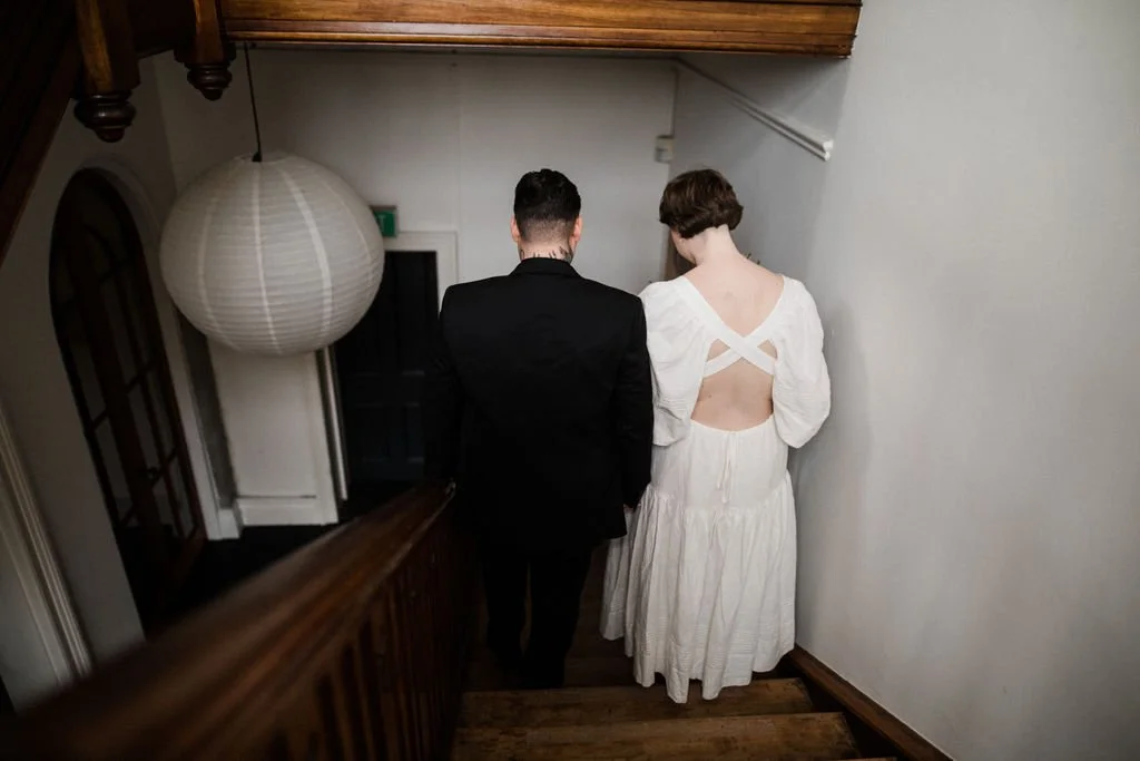 A man and a woman walking down a staircase from behind, with the woman in a white dress with an open back and the man in a black suit.