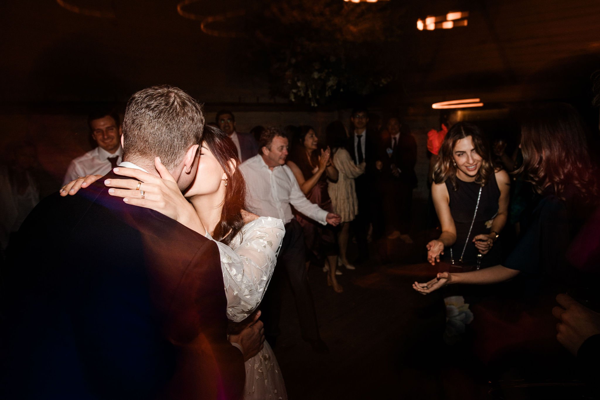 A couple kissing on a dance floor at a celebration or party, with friends and guests dancing and cheering in the background.