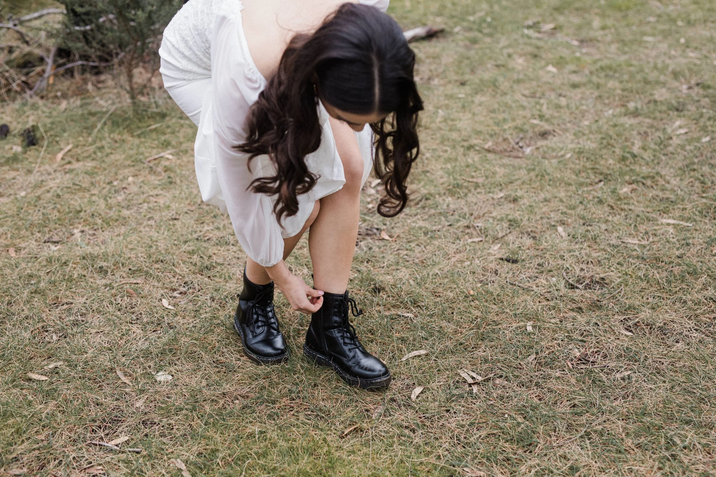 A woman wearing a white dress and black combat boots is outdoors, bending down to tie her shoelace on her left foot, on a grassy area with some scattered dry leaves.