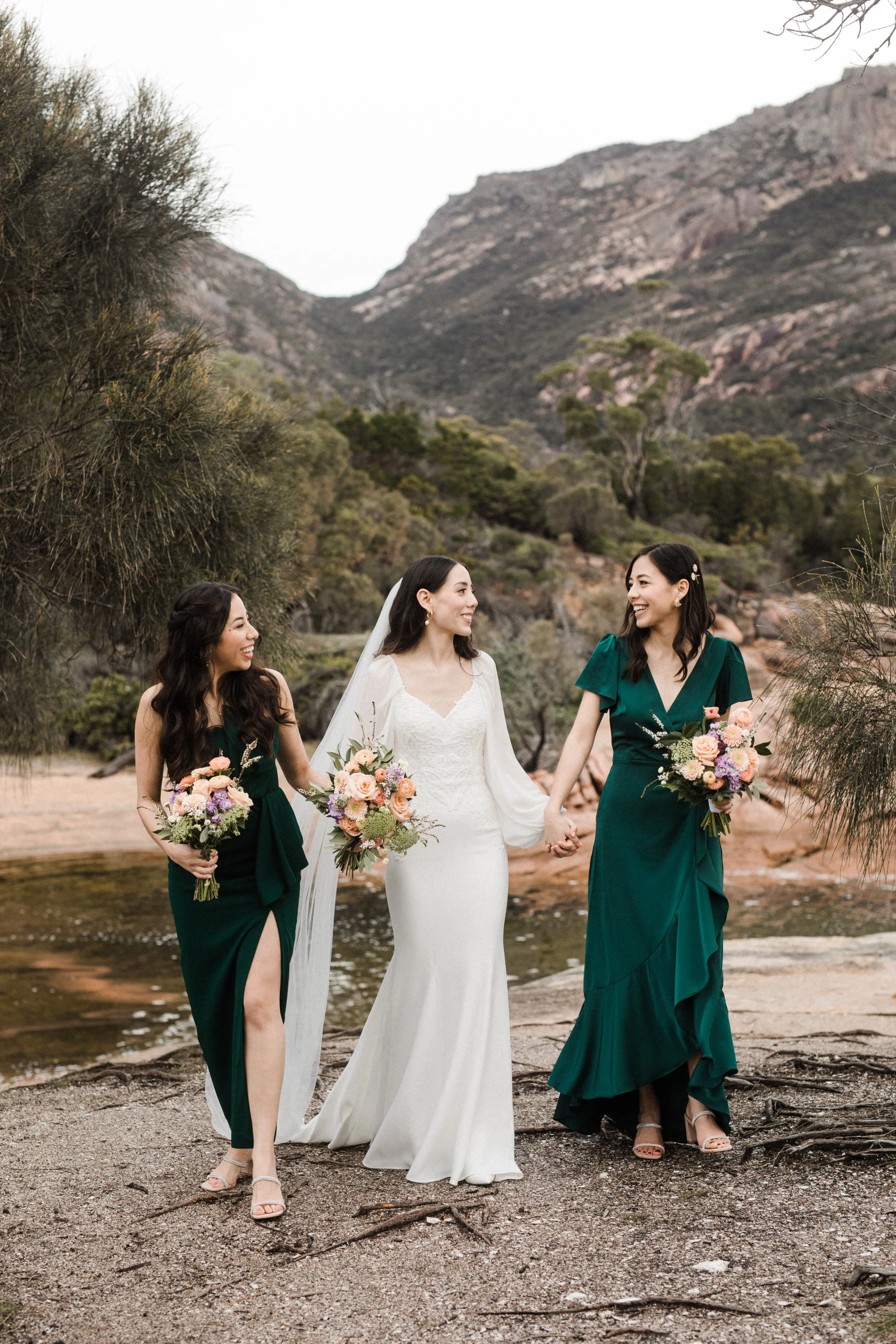 A bride in a white wedding dress holding hands with two bridesmaids in green dresses, all holding bouquets, walking outdoors near a water body with mountains in the background.