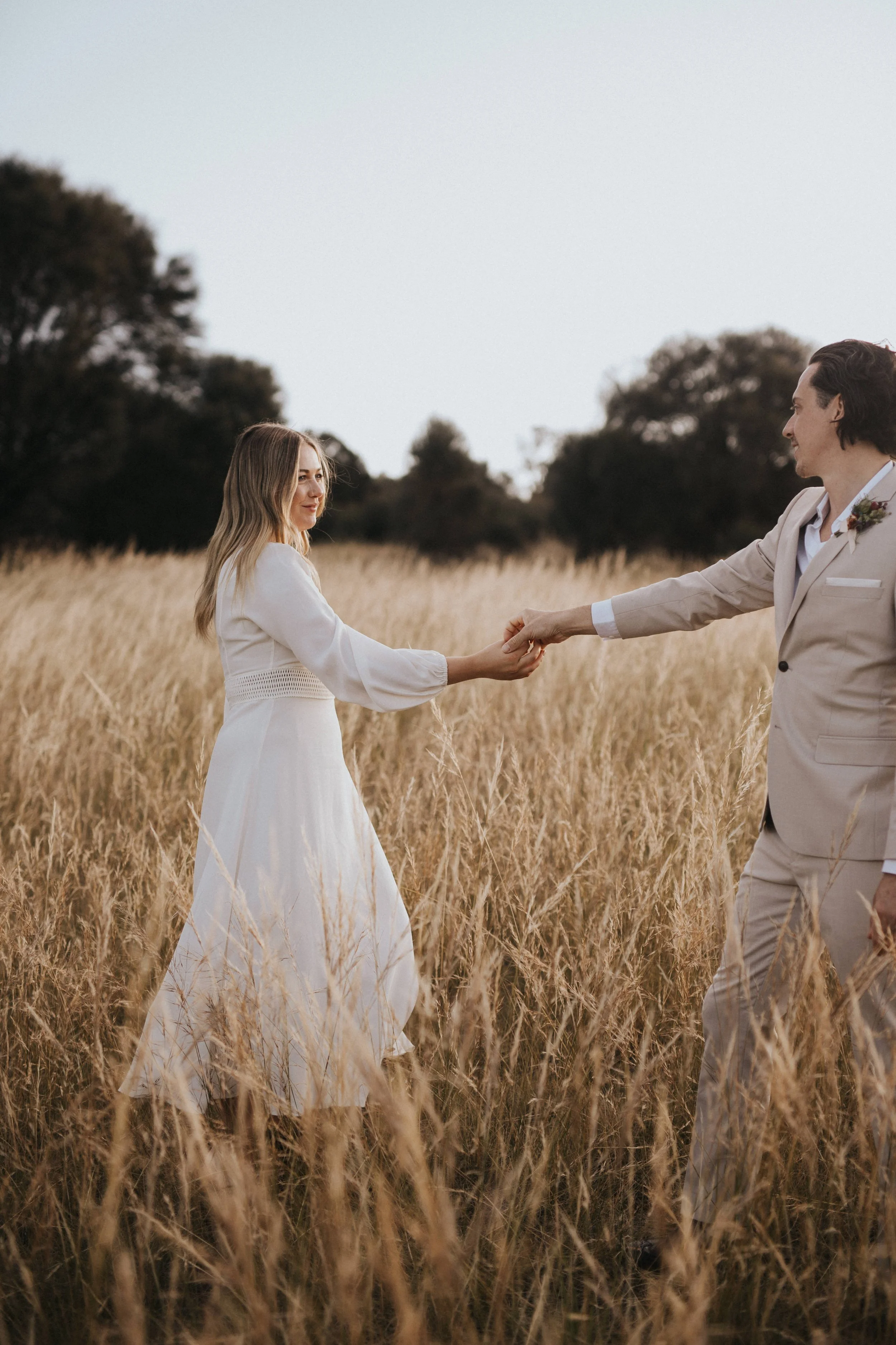 A couple holding hands in a field of tall, golden grass, with trees in the background during sunset or dusk.
