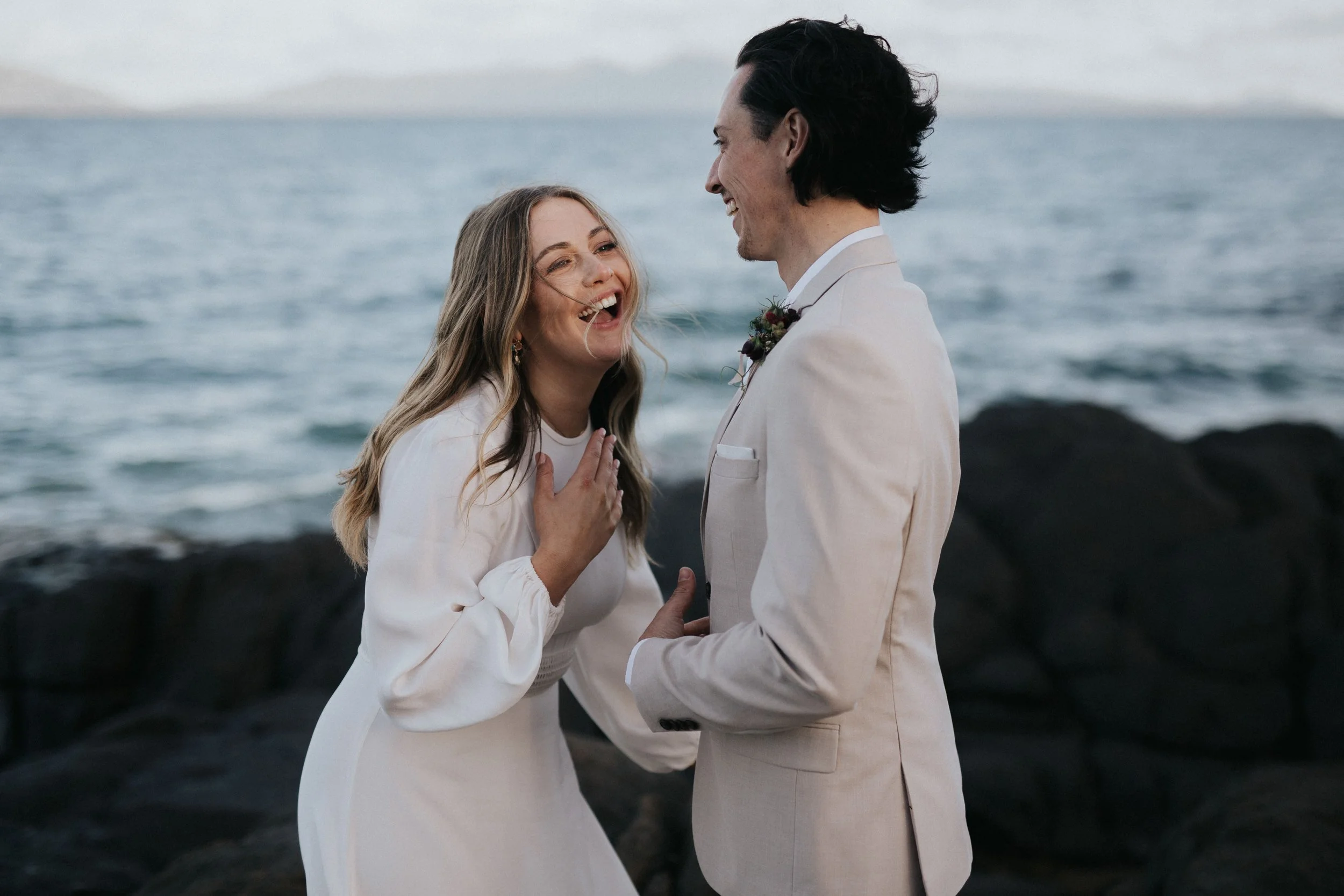 A couple in wedding attire standing by the ocean, smiling and laughing, with rocks and water in the background.