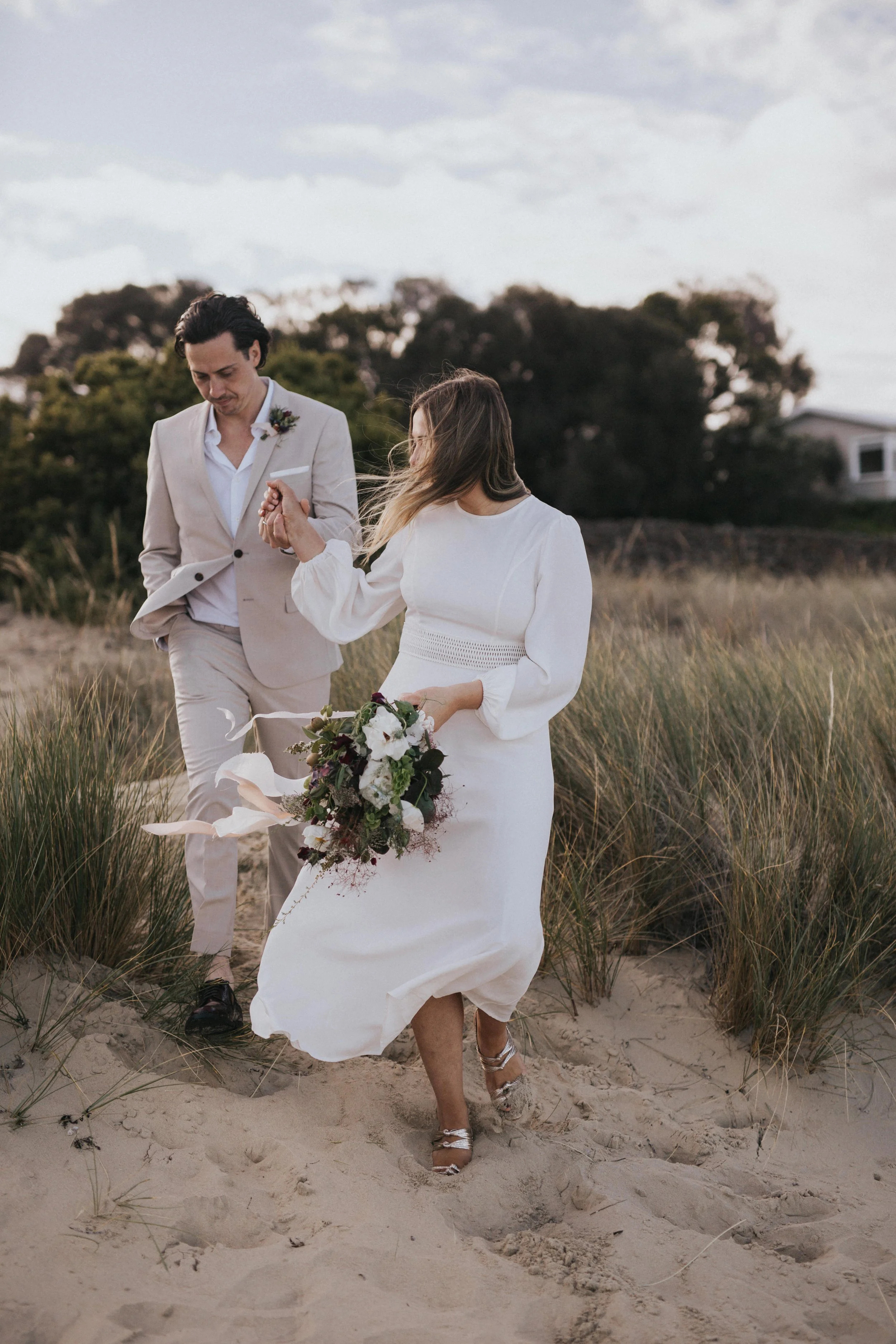 A couple walking on a sandy beach, holding hands, with the woman holding a bouquet of flowers and wearing a white dress, and the man dressed in a light-colored suit.