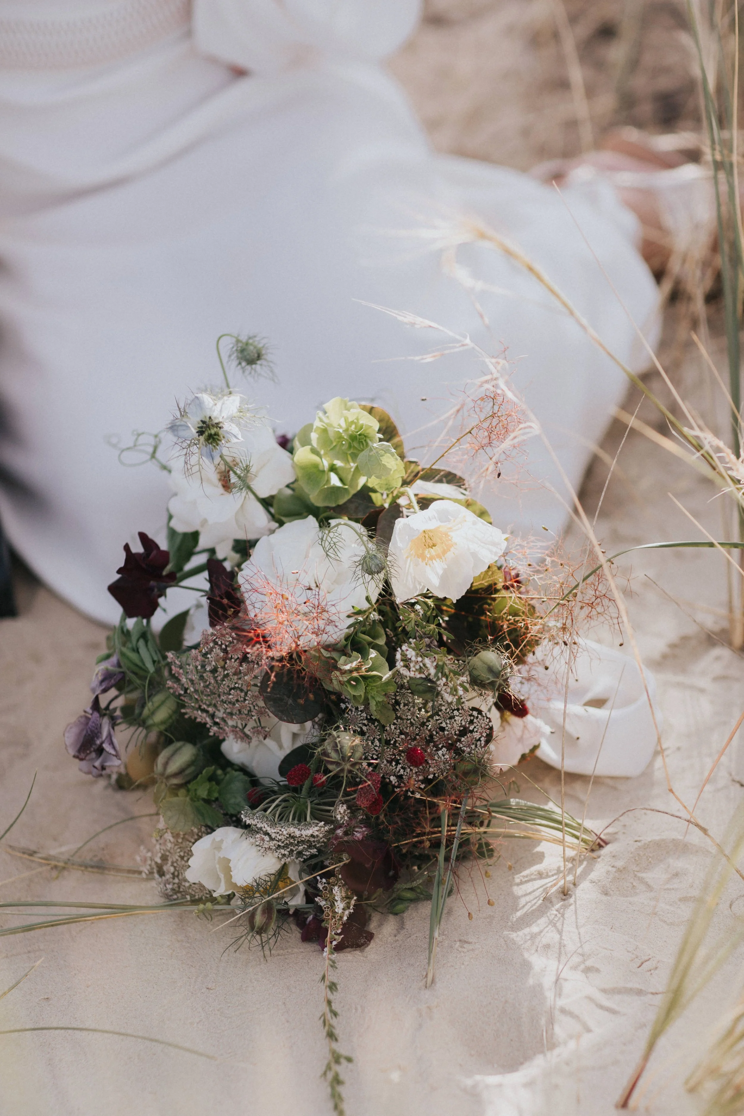 A bouquet of flowers resting on sandy ground, with a white cloth and beach grass in the background.