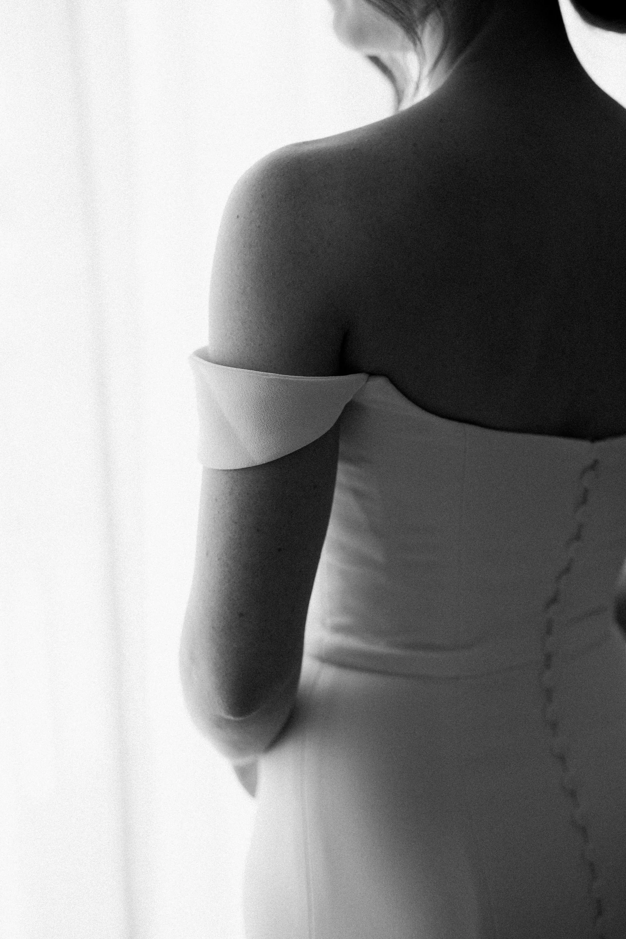 Close-up black and white photo of a woman in a strapless dress, with one shoulder uncovered, standing near a window with bright light behind her.