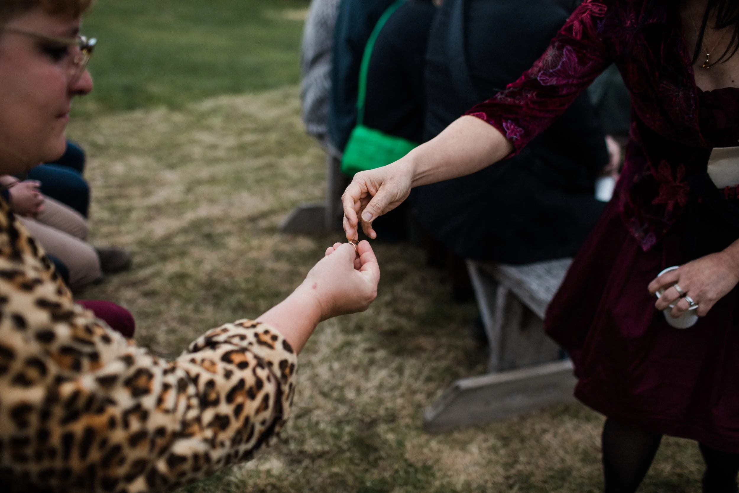 A person in a leopard print coat is exchanging rings with a woman dressed in a dark red velvet outfit outdoors, with other people and benches visible in the background.