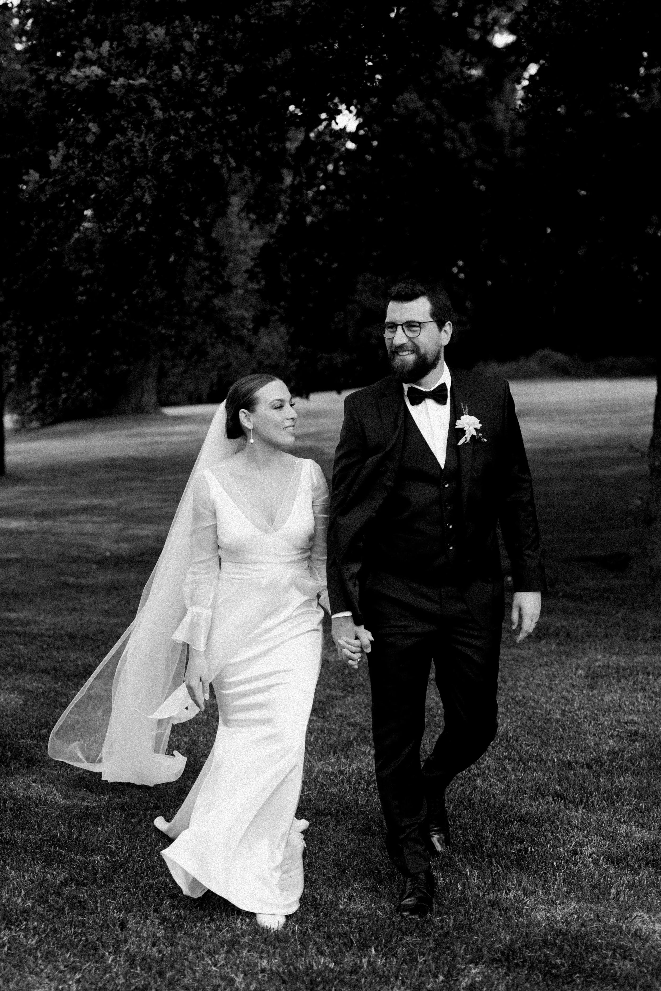 A bride and groom walking hand in hand outdoors, smiling, with trees in the background, black and white photograph at Quamby Estate, Tasmania.